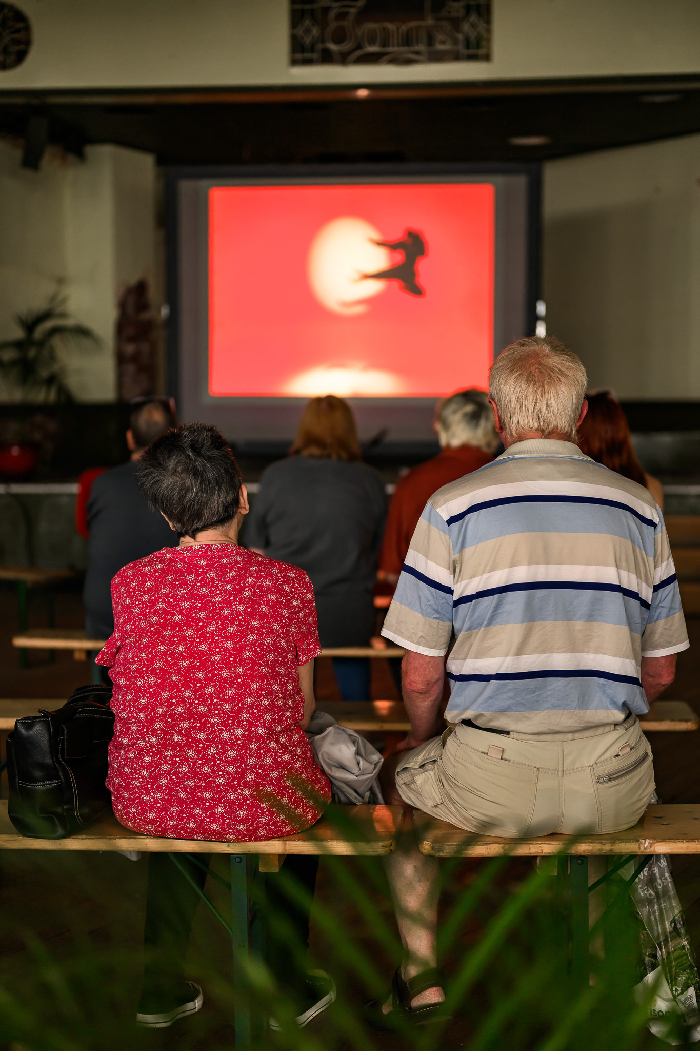 A group of people sits on benches indoors, facing a screen displaying a red background with a silhouette of a person jumping in front of a white circle, possibly representing the moon.