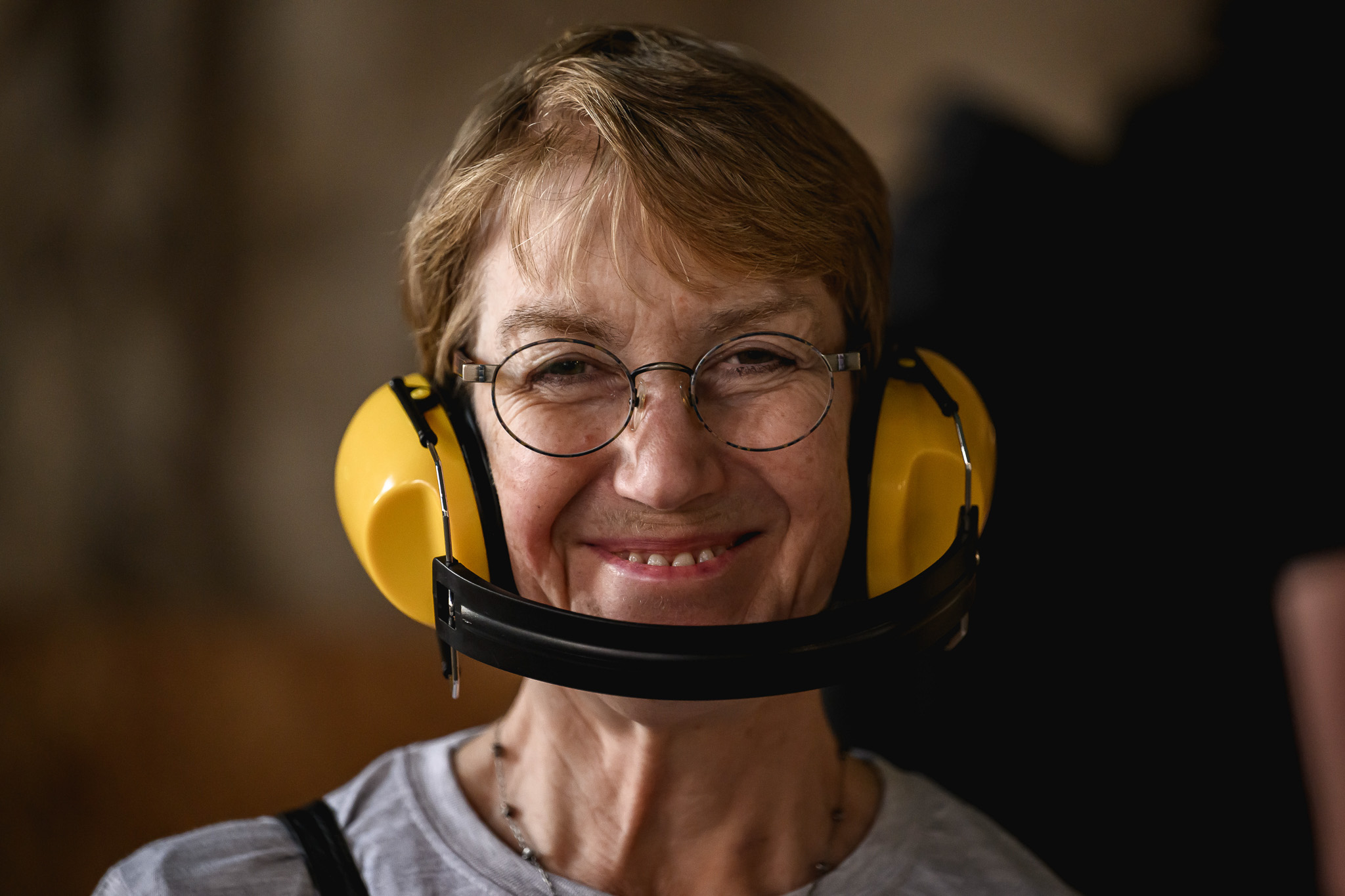 An older woman smiles at the camera, wearing round glasses and yellow ear protection headphones around her neck. She has short, light brown hair and is dressed casually. The background is softly blurred.