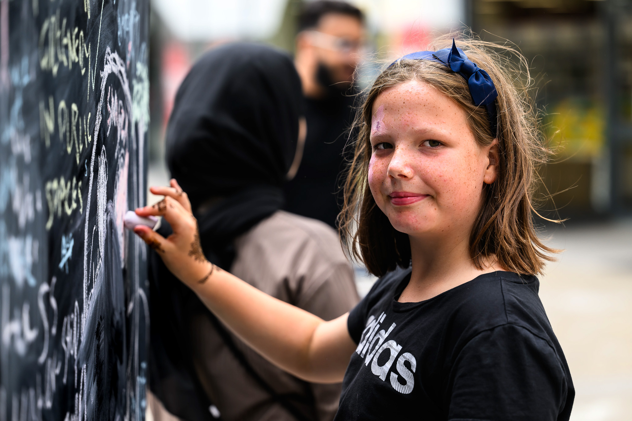 A young girl with brown hair and a blue bow smiles at the camera while writing on a blackboard. Another person in a black hijab stands beside her, also drawing on the board. The scene appears to be outdoors.