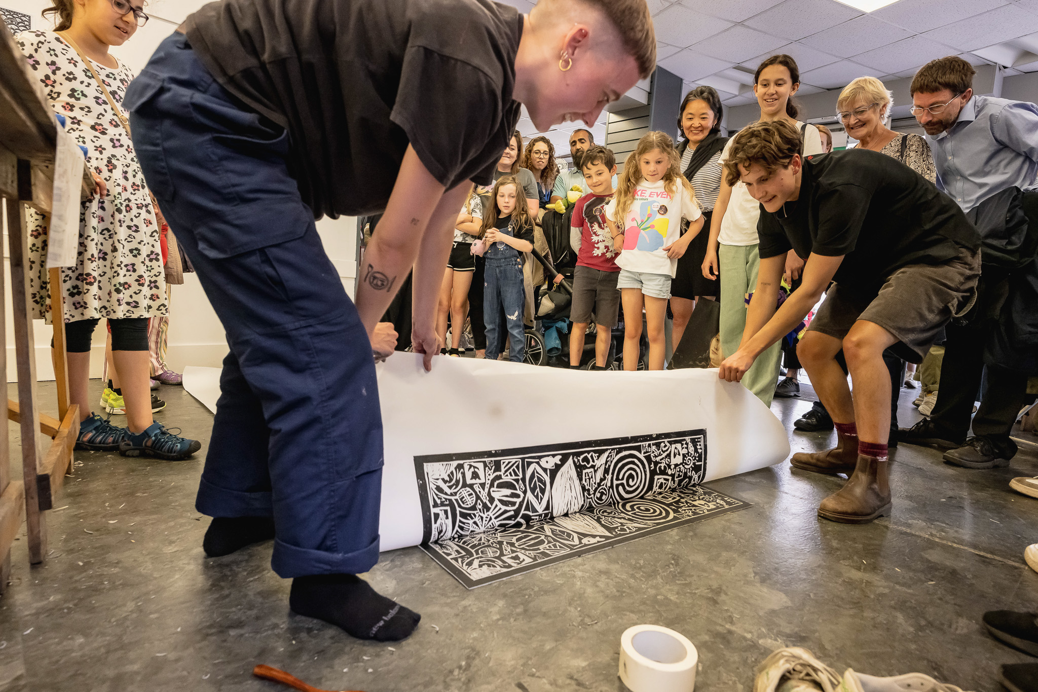 Two people lift a large sheet of paper to reveal a black and white print on the floor, whilst a group of smiling onlookers, including children and adults, watch the art demonstration in a bright, indoor space.