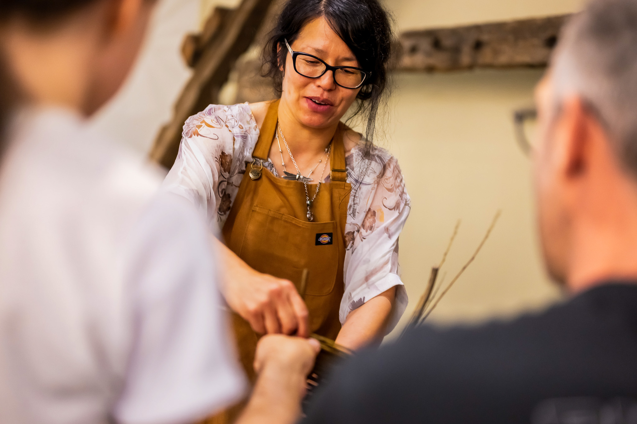 A woman wearing glasses and a brown apron demonstrates a craft or activity to two people, whose backs are to the camera, in a warm indoor setting.