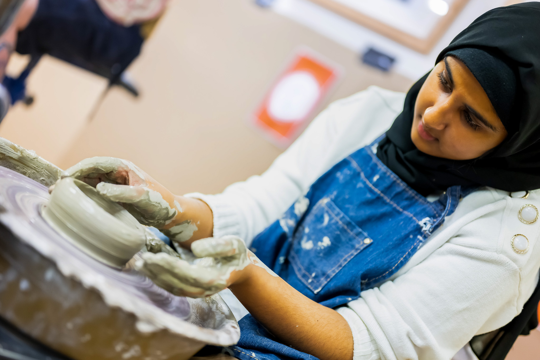 A woman wearing a black hijab and blue apron shapes clay on a potter’s wheel, focussing intently as she uses both hands to mould the spinning clay.