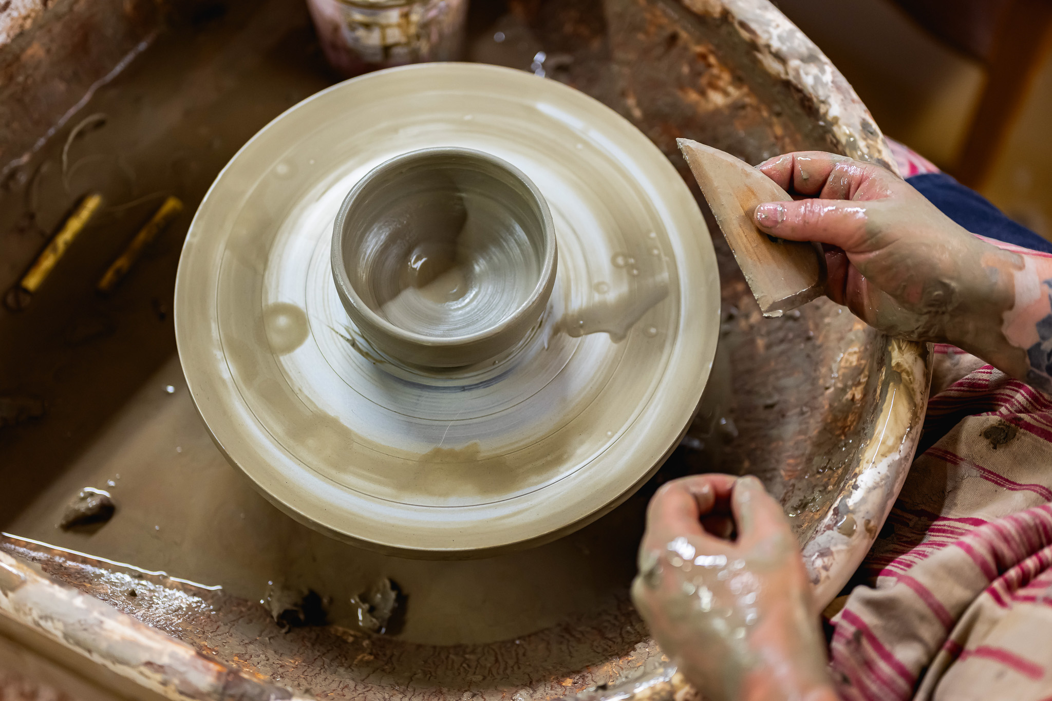 A person shapes a wet clay pot on a spinning potter's wheel, using a tool with one hand and supporting the clay with the other. Their hands are covered in wet clay.