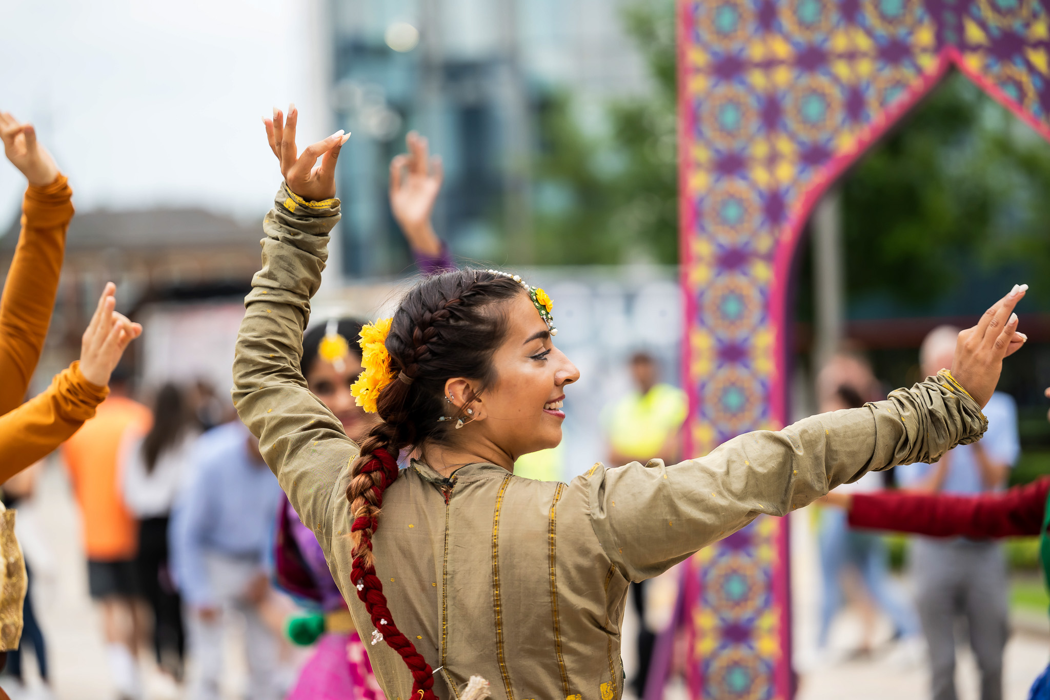 A woman in traditional attire dances outdoors with others, her arms raised and decorated with flowers. Colourful patterns and blurred people are visible in the background.