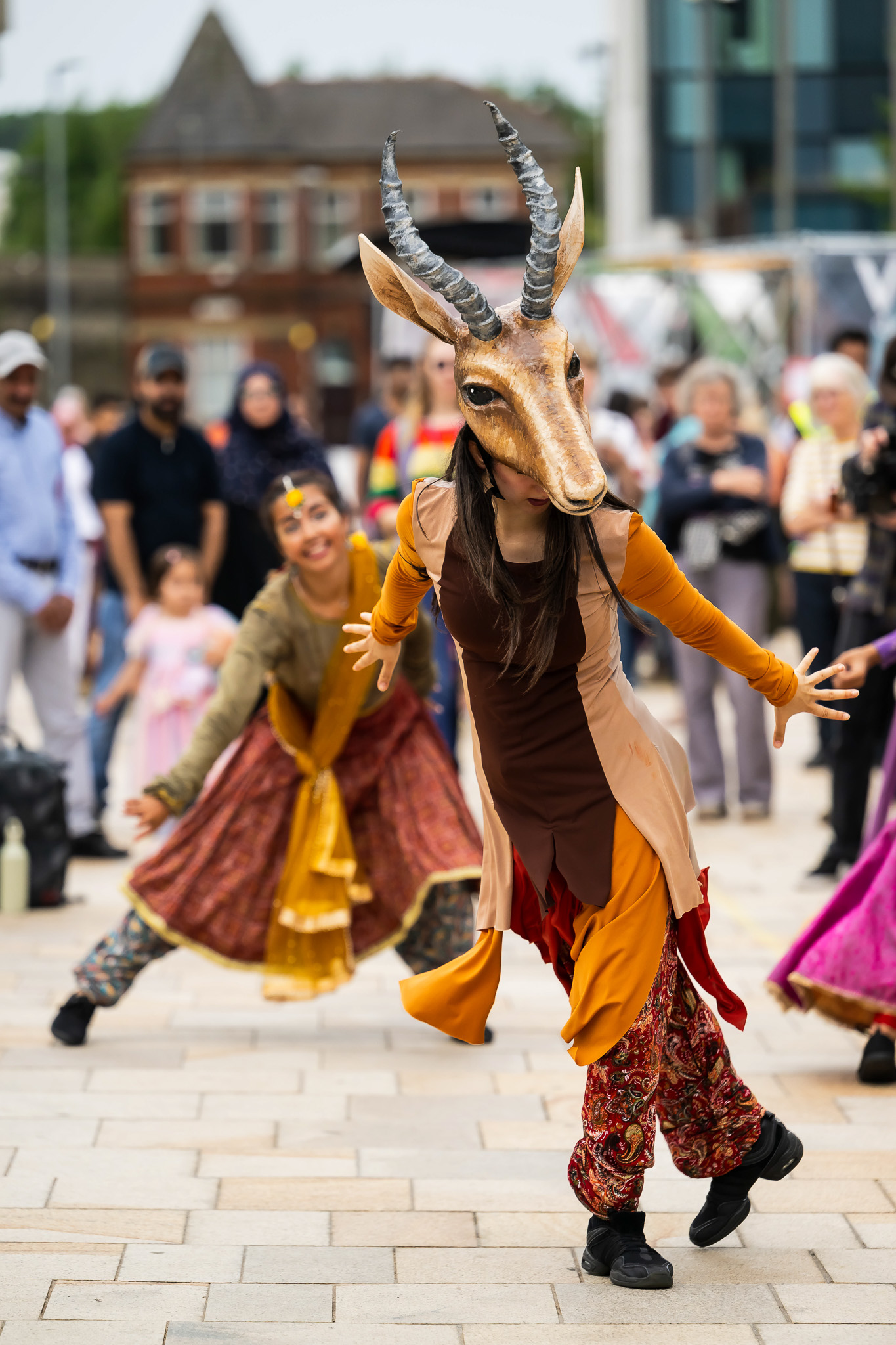 A performer in a gazelle mask and colourful costume dances energetically outdoors, with another dancer behind her. A crowd watches in the background on a paved plaza.
