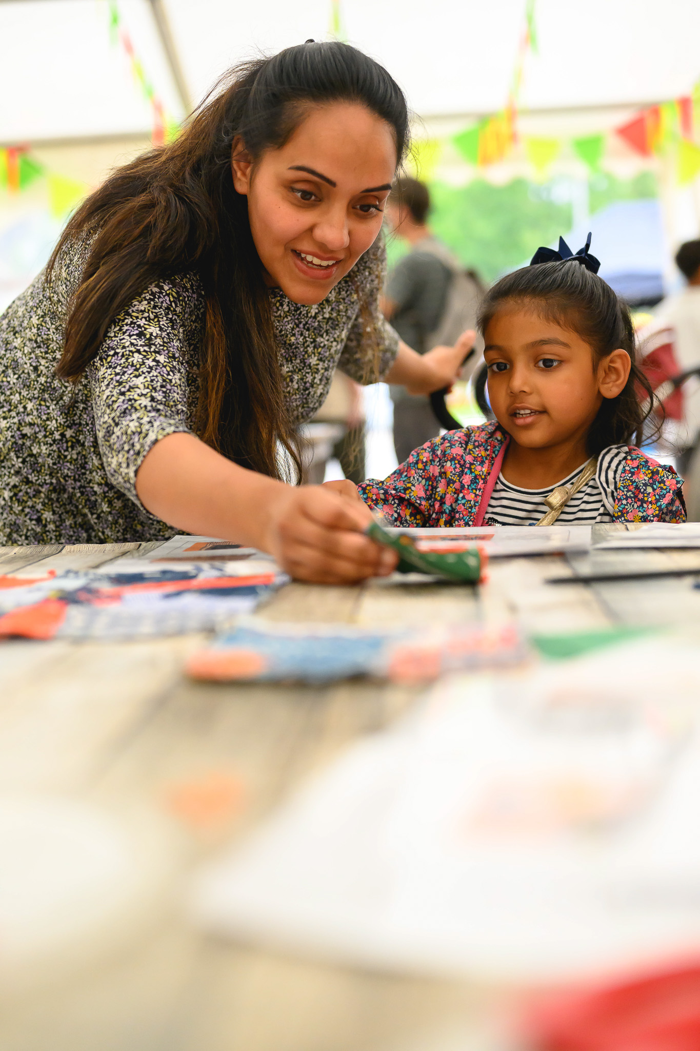 A woman and a young girl sit at a table covered with craft supplies. The woman smiles as she shows the girl something, whilst the girl looks on with curiosity. Colourful decorations hang in the background.