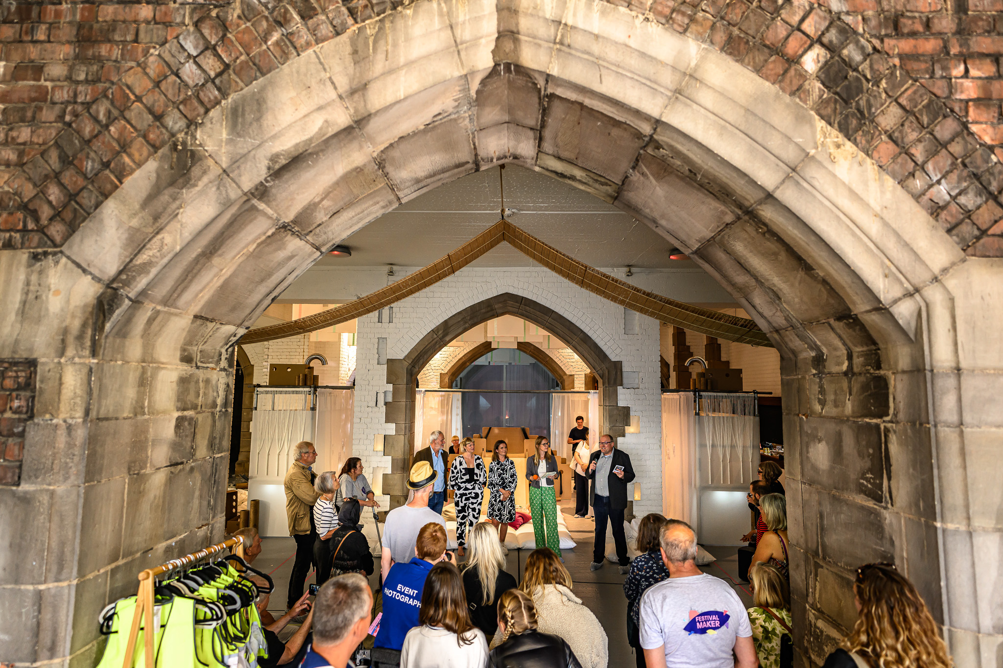 A group of people gathers inside a vaulted, arched brick structure with gothic-style architecture, listening to speakers at the front of the room. Clothing hangs on a rail to the left.