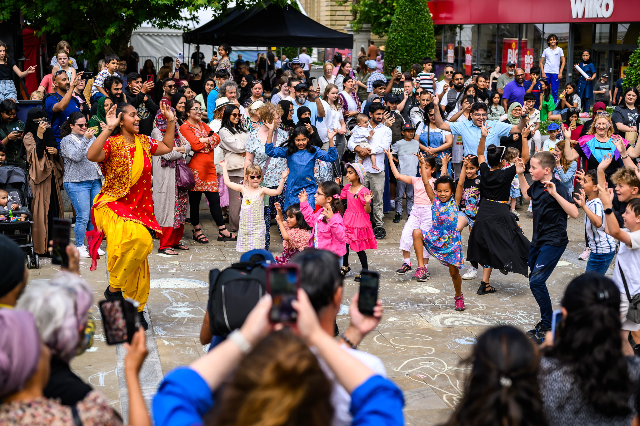 A large crowd, including children and adults, watch and join a woman in leading a lively outdoor dance on a chalk-decorated pavement. Many people are smiling and taking photos, enjoying the festive atmosphere.