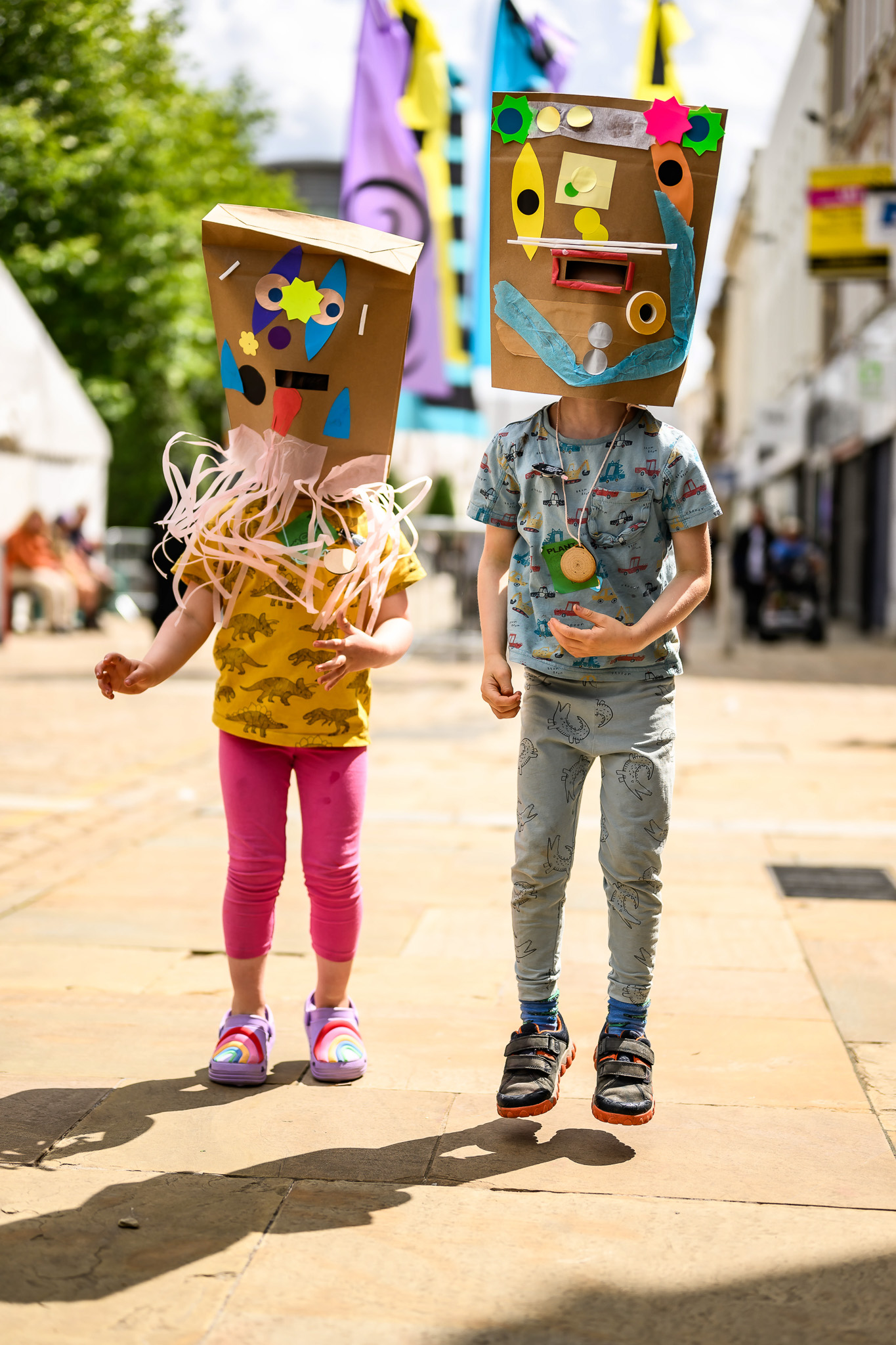 Two children wearing colourful, decorated cardboard box masks jump in the air on a sunny street, with vibrant flags and tents visible in the background.