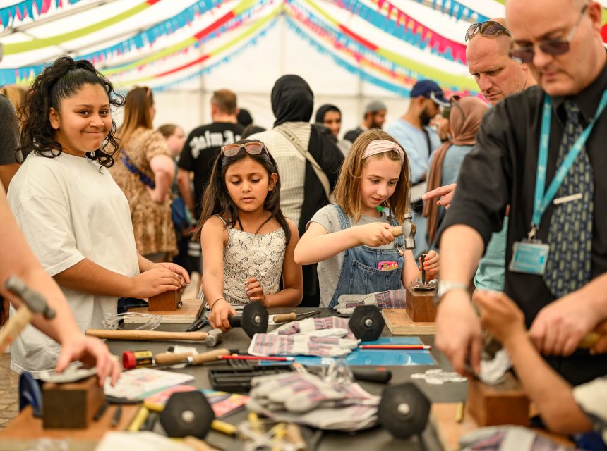 Children and adults participate in a hands-on craft activity under a colourful, decorated marquee. People are using tools and working on creative projects at crowded tables, with a lively group in the background.