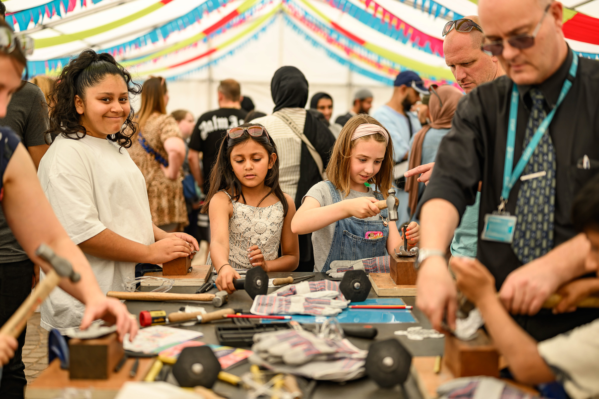 Children and adults participate in a hands-on craft activity under a colourful, decorated marquee. People are using tools and working on creative projects at crowded tables, with a lively group in the background.