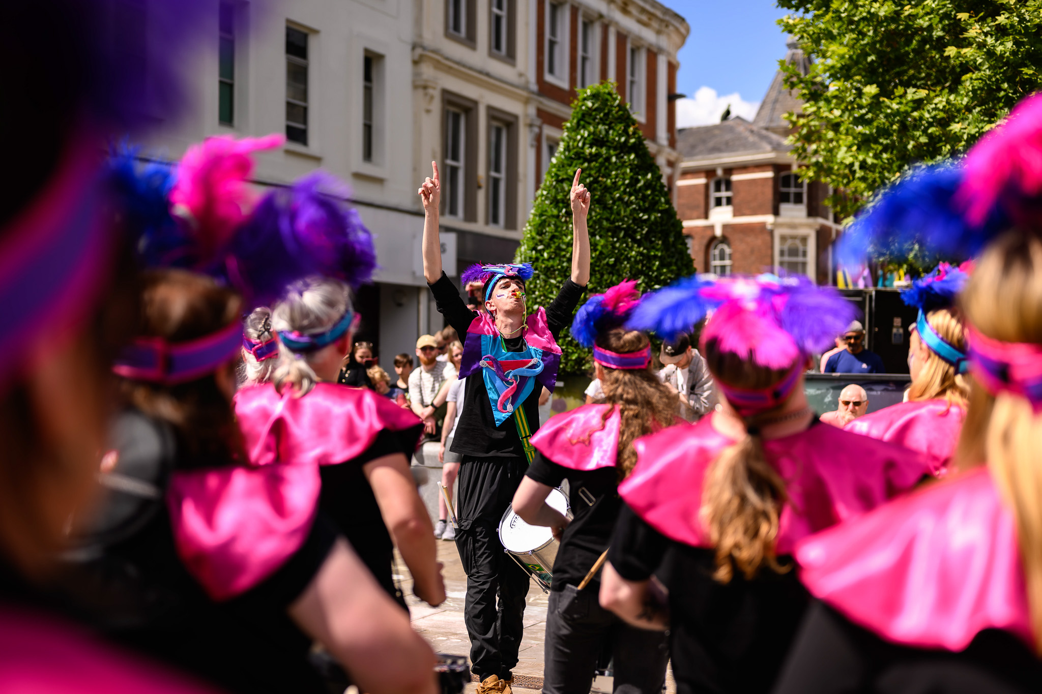 A group of people in vibrant costumes with pink and purple feathers perform outside, led by a person in the centre raising both hands, as onlookers watch on a sunny day.
