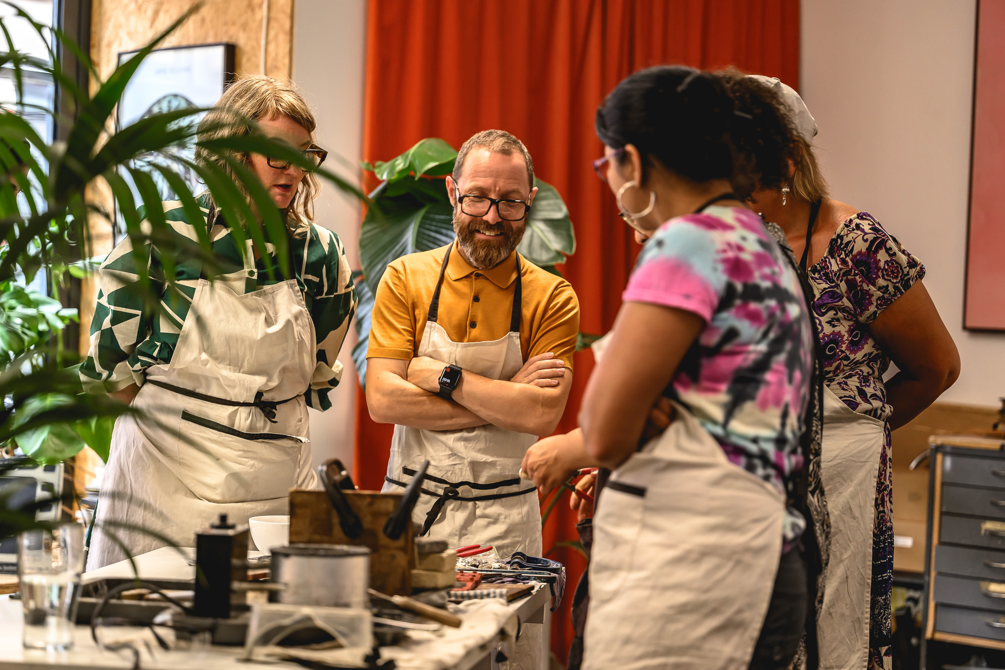 A group of four adults wearing aprons stand around a worktable with tools and materials, engaged in a creative or craft activity in a brightly lit indoor space with plants and a red curtain.