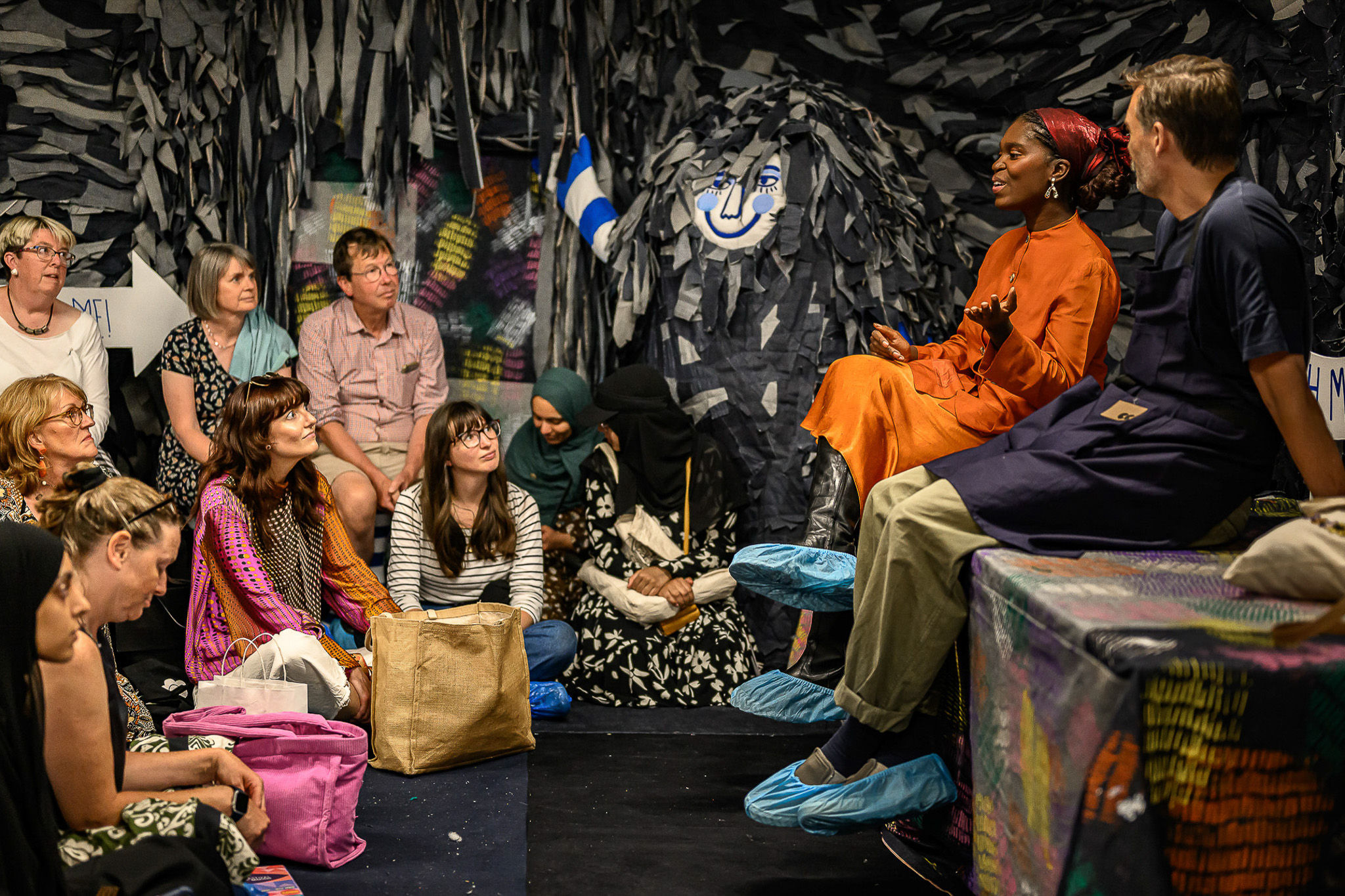 A diverse group of people sit attentively on the floor in a decorated room, listening to two speakers. One woman in an orange dress gestures while speaking. Everyone wears blue shoe covers. The room is filled with dark, hanging decorations.