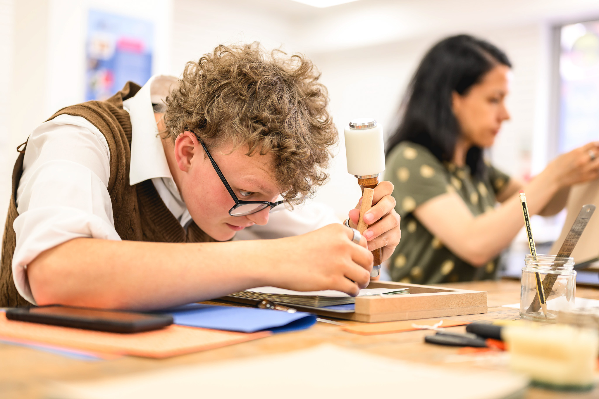 A young person with curly hair and glasses focuses on crafting with a mallet and tool at a table, while an adult works on a separate project in the background. Art supplies are scattered on the table.