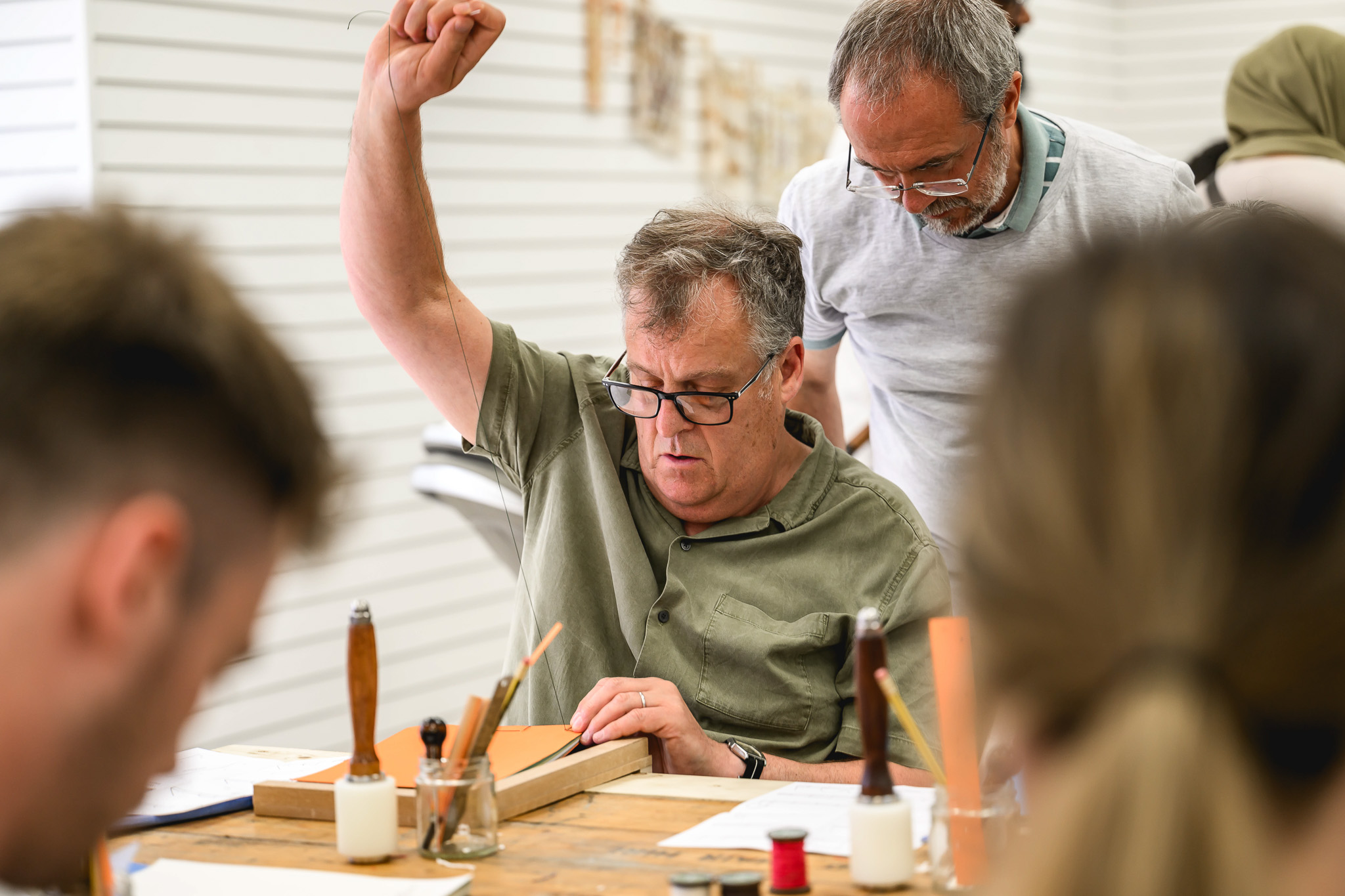 A man with glasses raises his hand whilst sitting at a workshop table with tools and materials, as another man stands beside him, observing or assisting. Other people are blurred in the foreground.