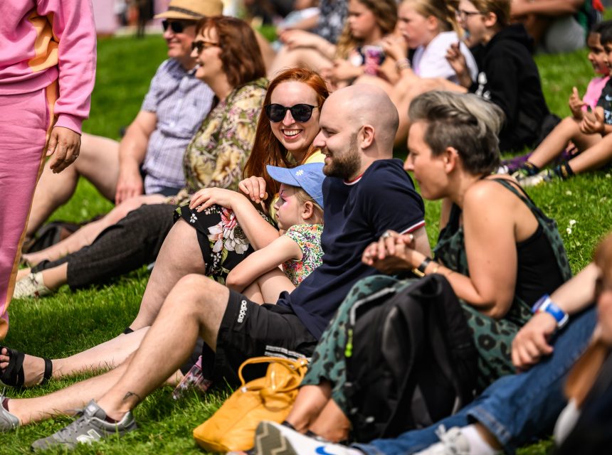 A group of people, including adults and children, sit together on a grassy hill in the sunshine, smiling and enjoying an outdoor event. Some wear sunglasses and hats, and rucksacks are visible on the grass.