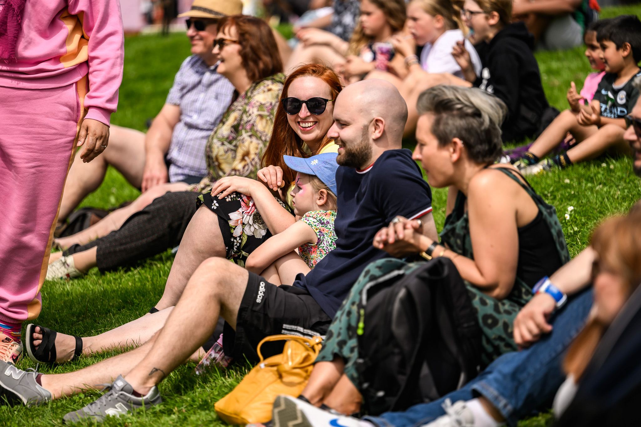 A group of people, including adults and children, sit together on a grassy hill in the sunshine, smiling and enjoying an outdoor event. Some wear sunglasses and hats, and rucksacks are visible on the grass.