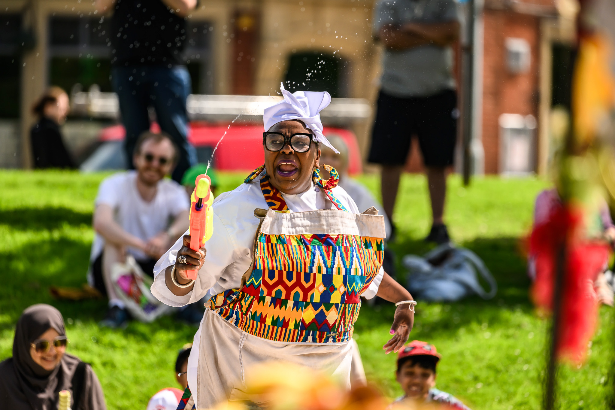 A joyful woman wearing colourful clothes and a pinafore sprays a water pistol outdoors, surrounded by smiling people sitting on grass on a sunny day.