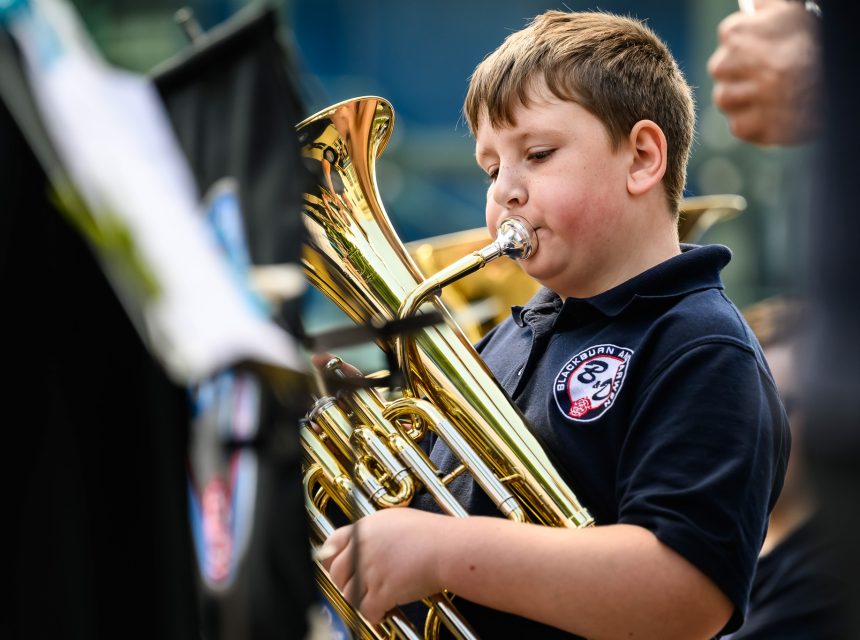 Young boy in a navy shirt plays a brass instrument in an outdoors performance.