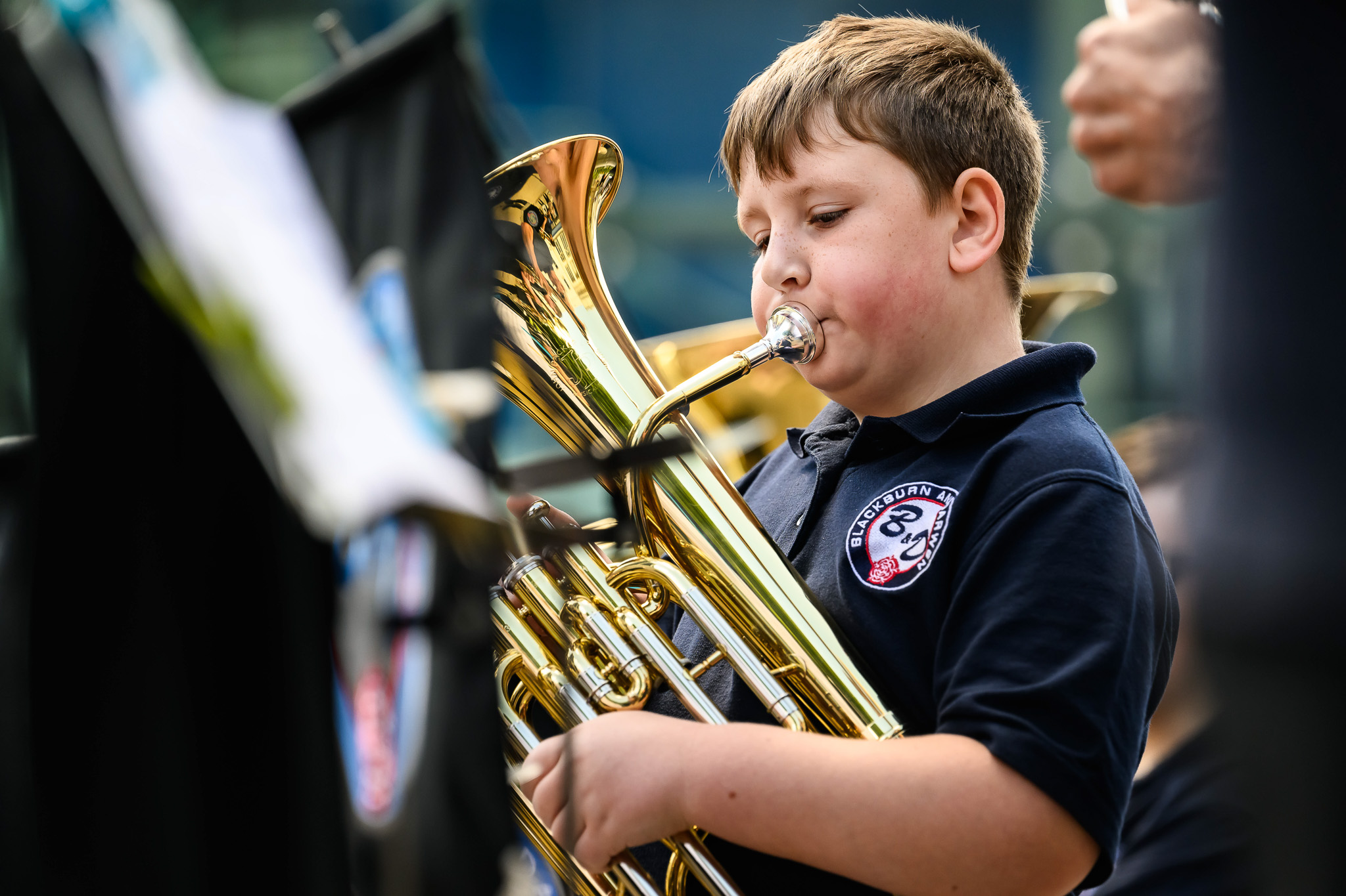 Young boy in a navy shirt plays a brass instrument in an outdoors performance.