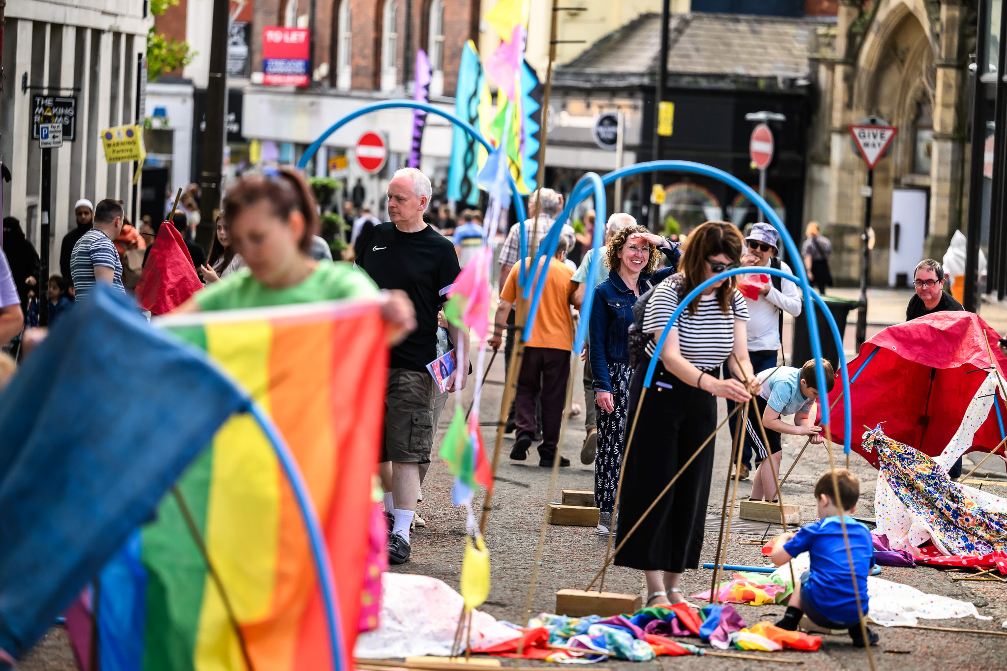 People of various ages gather on a city street, setting up colourful fabric structures and decorations. The atmosphere is lively and festive, with bright colours, flags, and playful activities.
