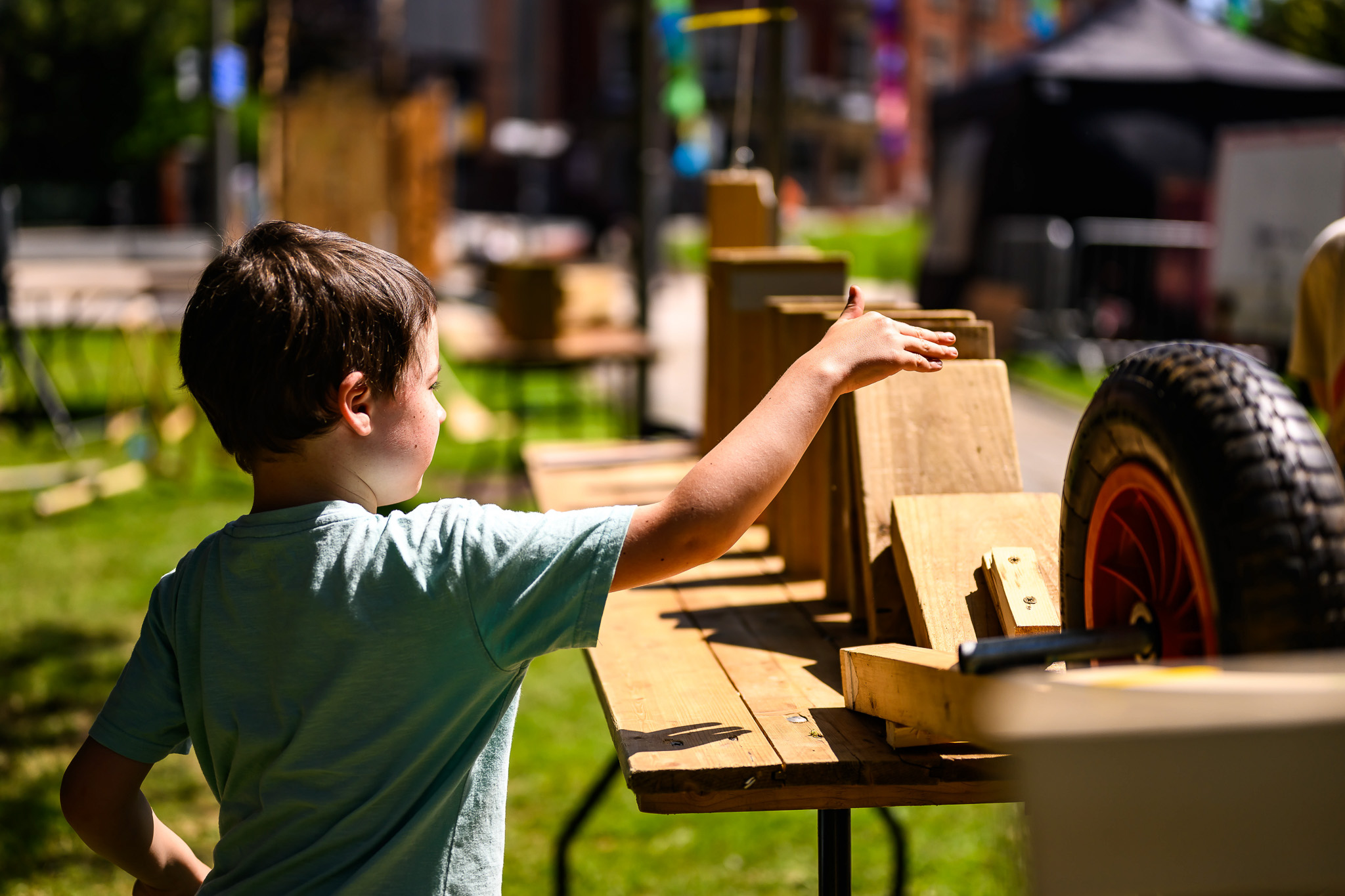 A young boy in a light blue shirt reaches out to touch wooden blocks and planks arranged on a table outdoors on a sunny day. A wheelbarrow tyre and other playground items are nearby.