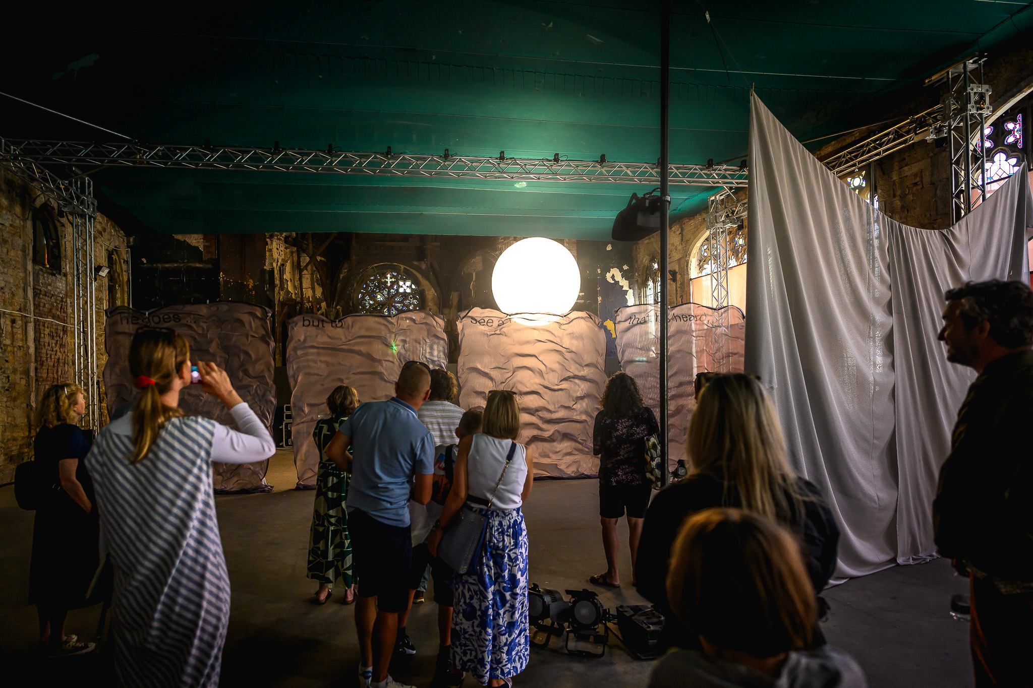 A group of people view an indoor art installation with a large illuminated sphere resembling the moon, set against textured backdrops and draped fabric under teal ceiling panels.