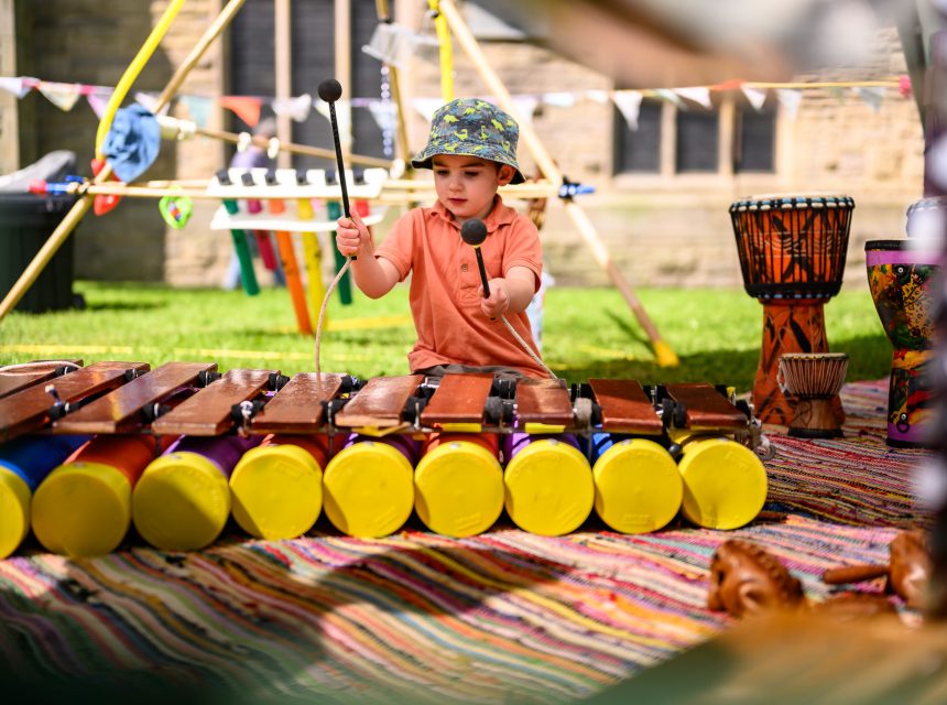 A young child wearing a colourful bucket hat and orange shirt plays a wooden xylophone outdoors, seated on a striped mat with percussion instruments around, at what appears to be a festival or event.
