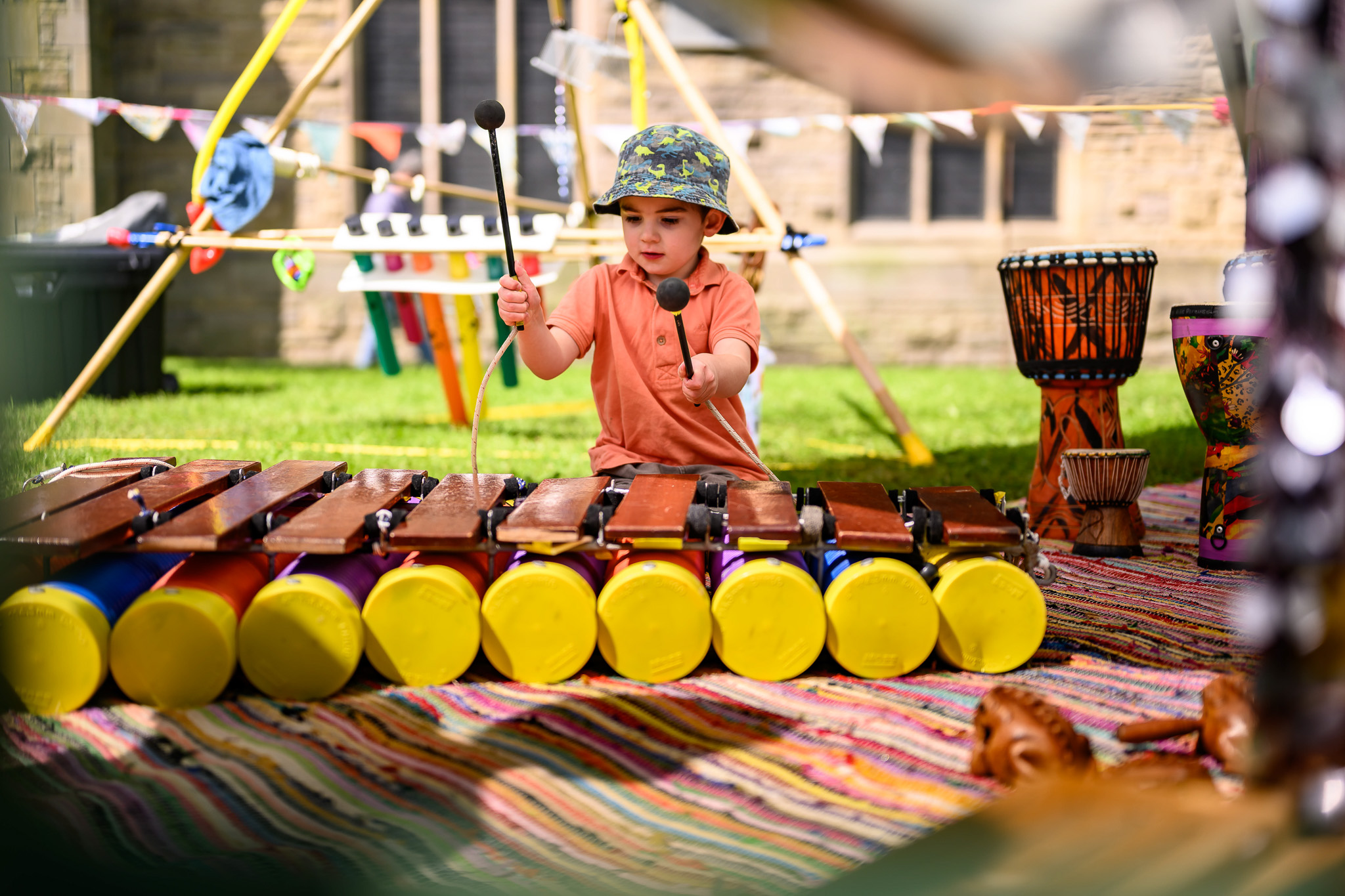A young child wearing a colourful bucket hat and orange shirt plays a wooden xylophone outdoors, seated on a striped mat with percussion instruments around, at what appears to be a festival or event.