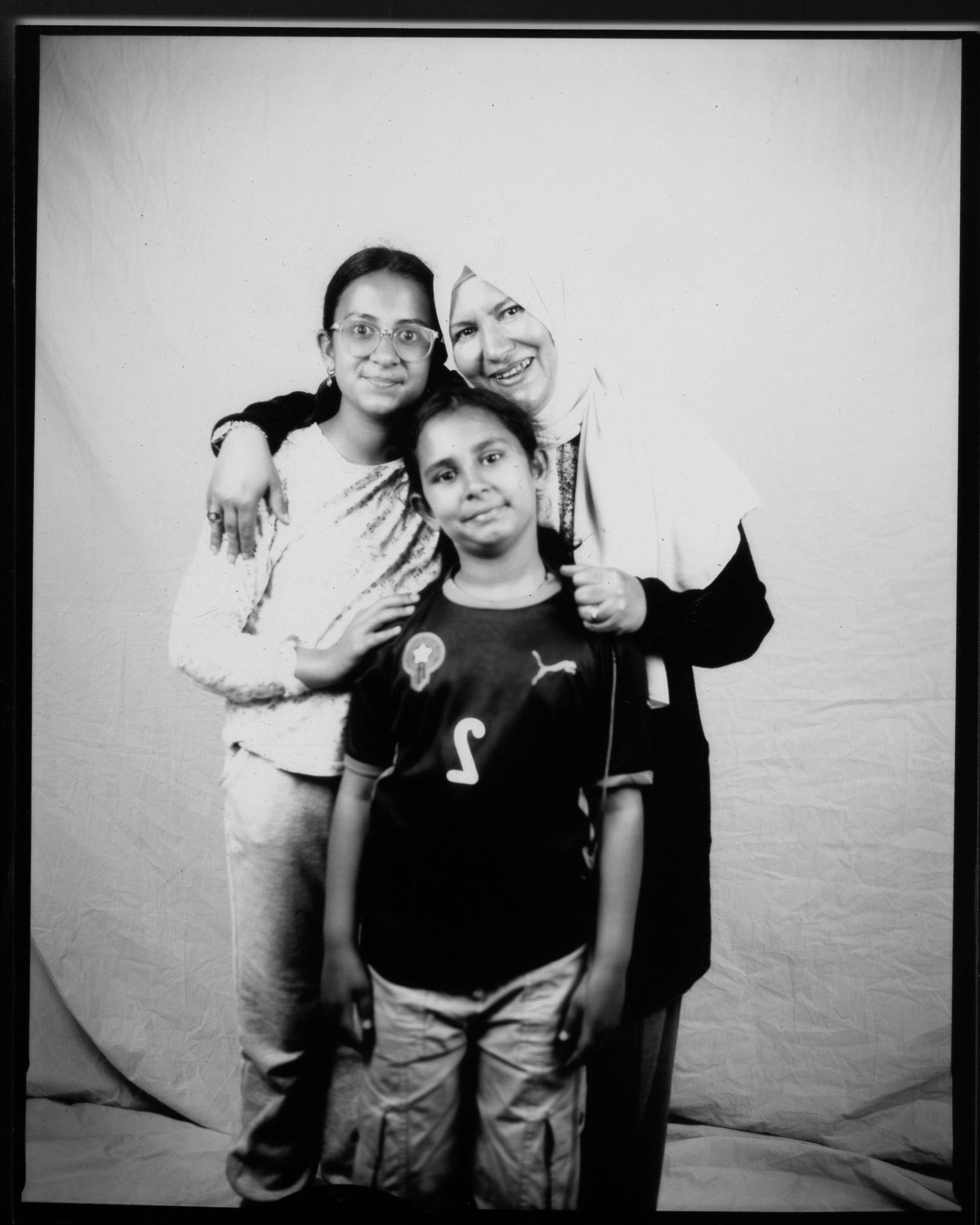 Black and white image of an adult with their arms around two children, all smiling in front of a white sheet.