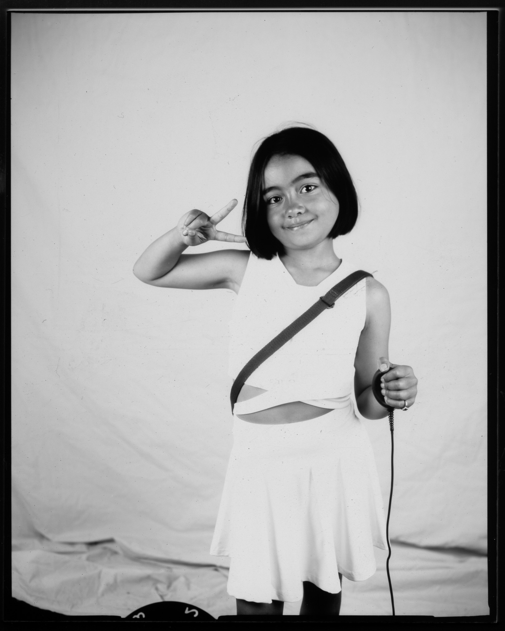 Black and white image of a child stood smiling with one hand in a peace sign. The other presses a remote, a wire trailing.