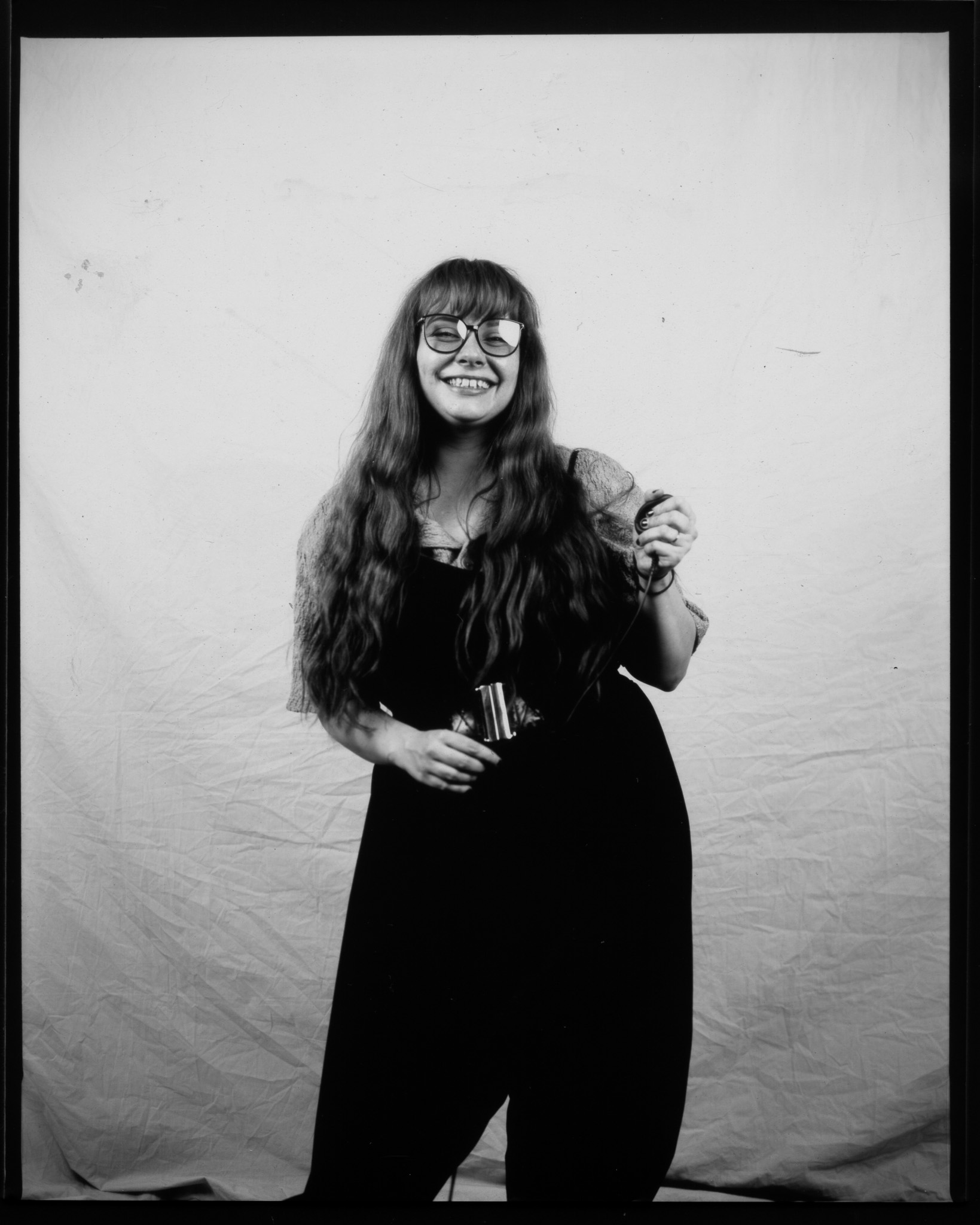 Black and white image of a smiling adult stood in front of a white sheet, pressing a remote shutter with a trailing wire.