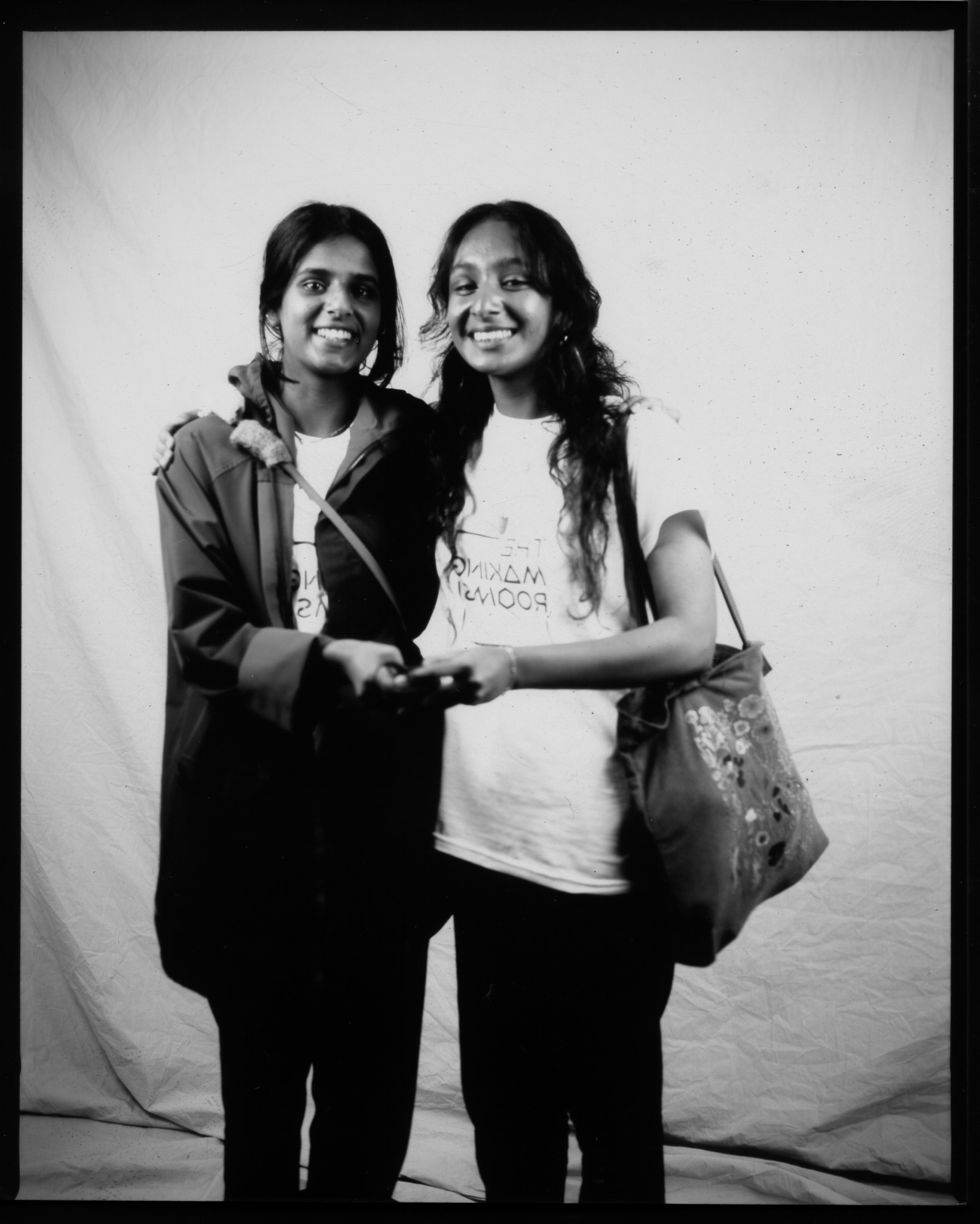 Black and white image of two young adults stood smiling in front of a white sheet. Both their hands pressing a remote.
