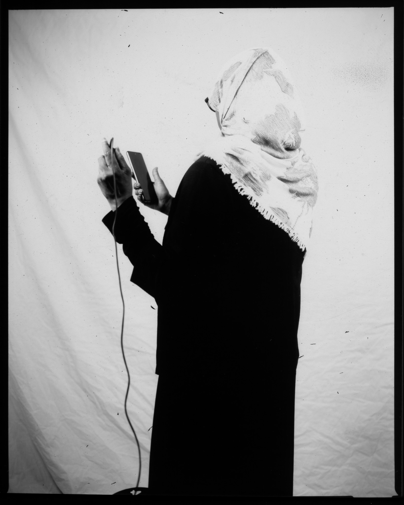 Black and white image of an adult turned away looking at a phone in front of a white sheet, pressing a remote with a wire.