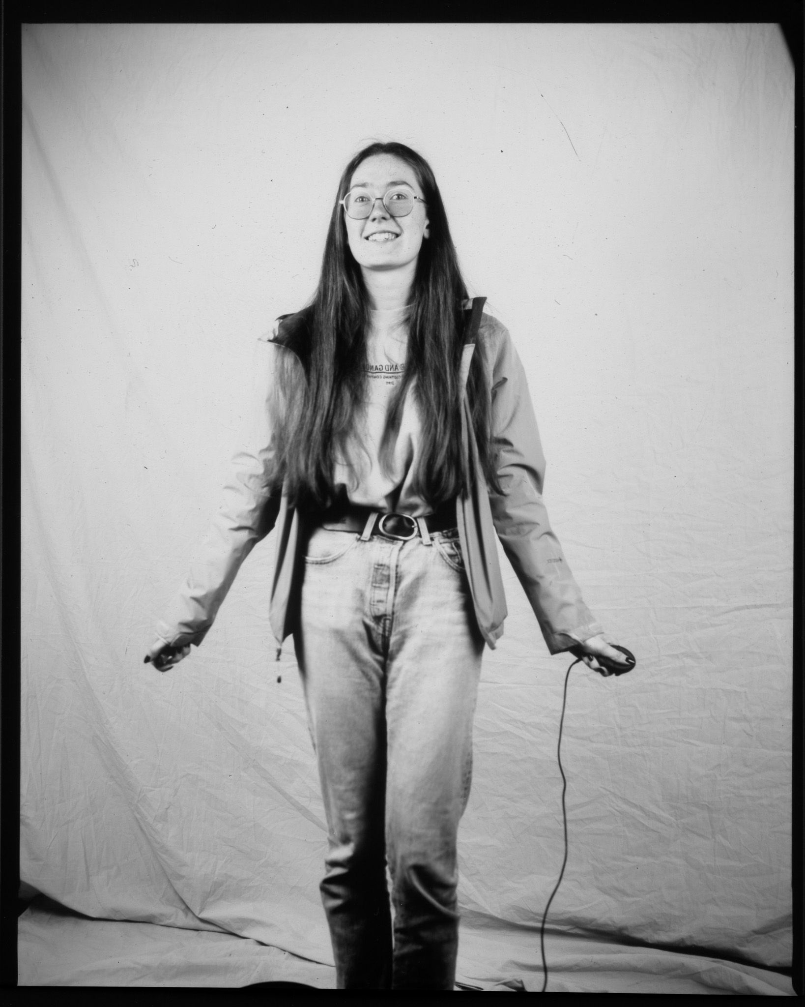 Black and white image of an adult smiling with arms out in front of a white sheet, pressing a remote with trailing wire.