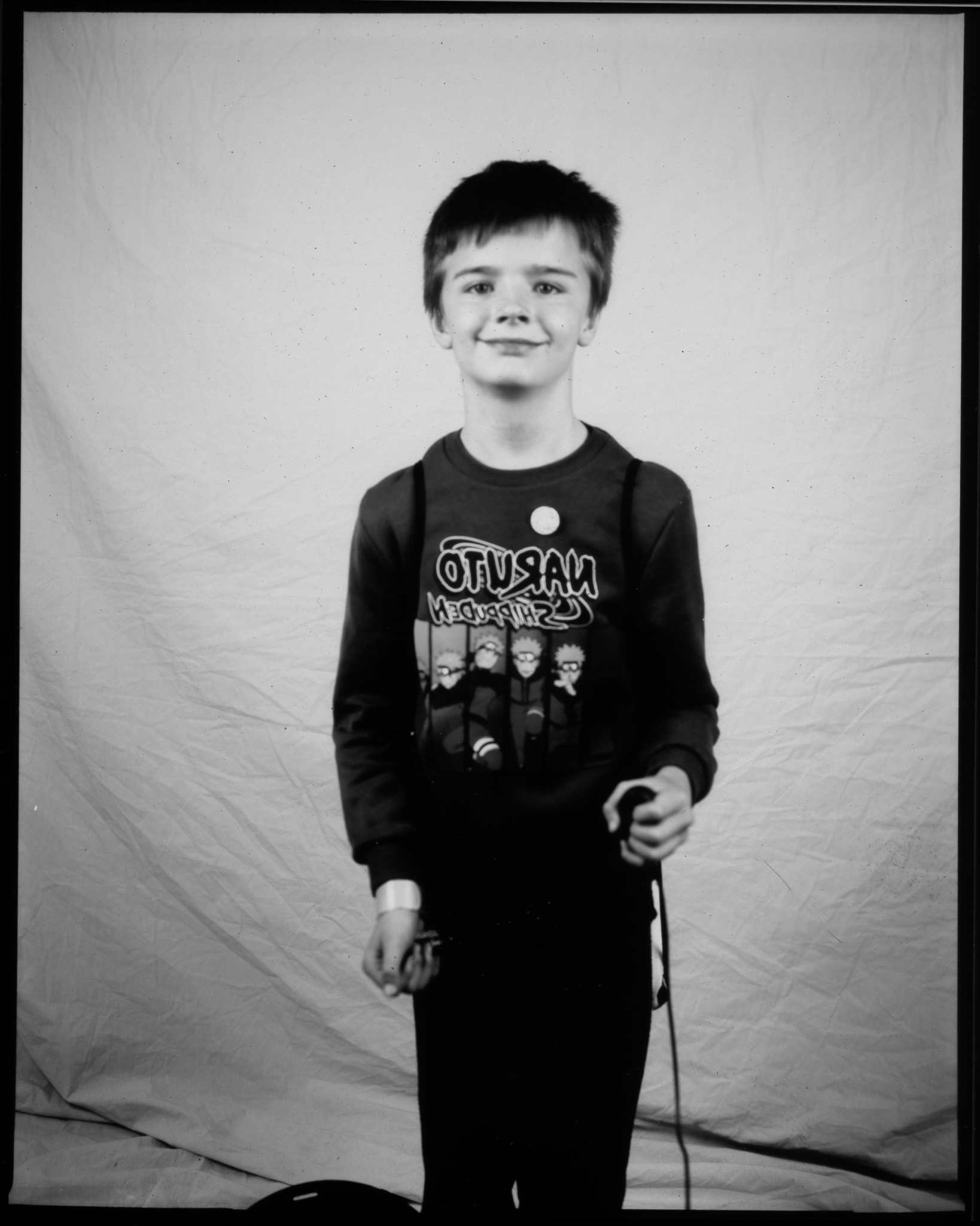 Black and white image of a child stood smiling in front of a white sheet, one hand pressing a remote with a wire trailing.