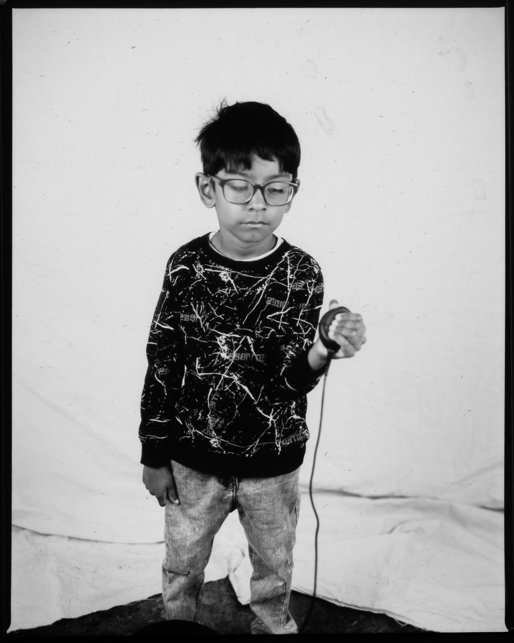 Black and white image of a child stood blinking in front of a white sheet, one hand pressing a remote with wire trailing.