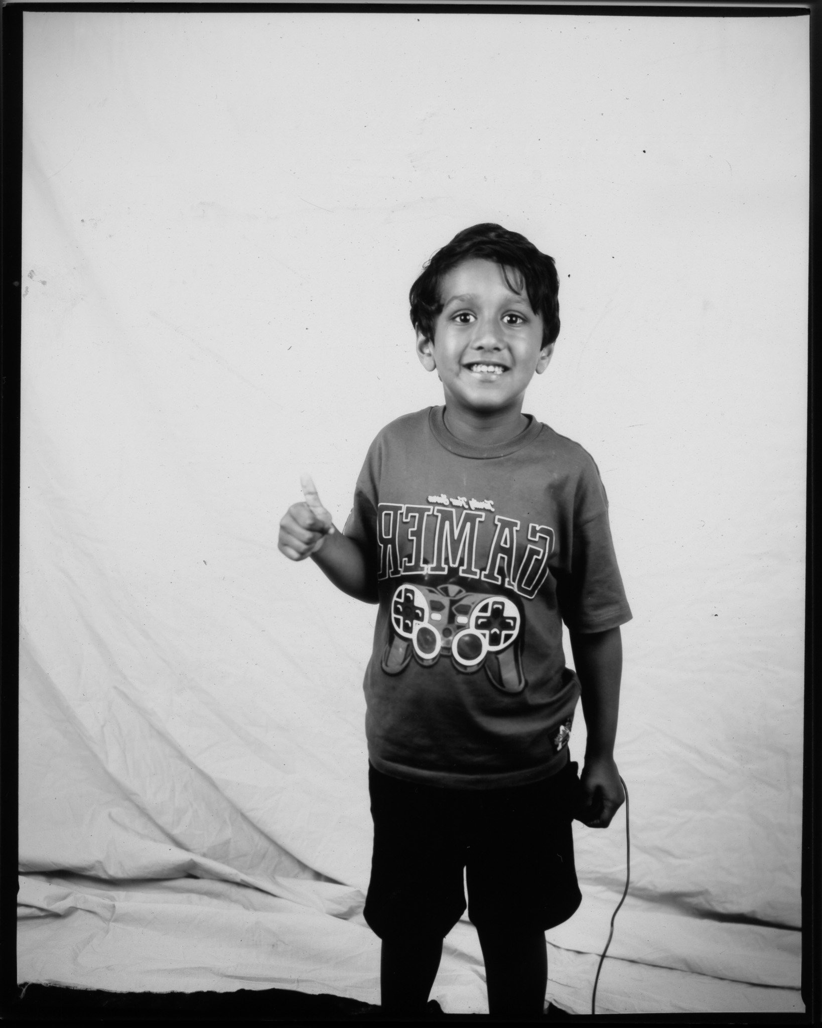 Black and white image of a child smiling with one hand in a thumbs up, the other pressing a remote with a trailing wire.