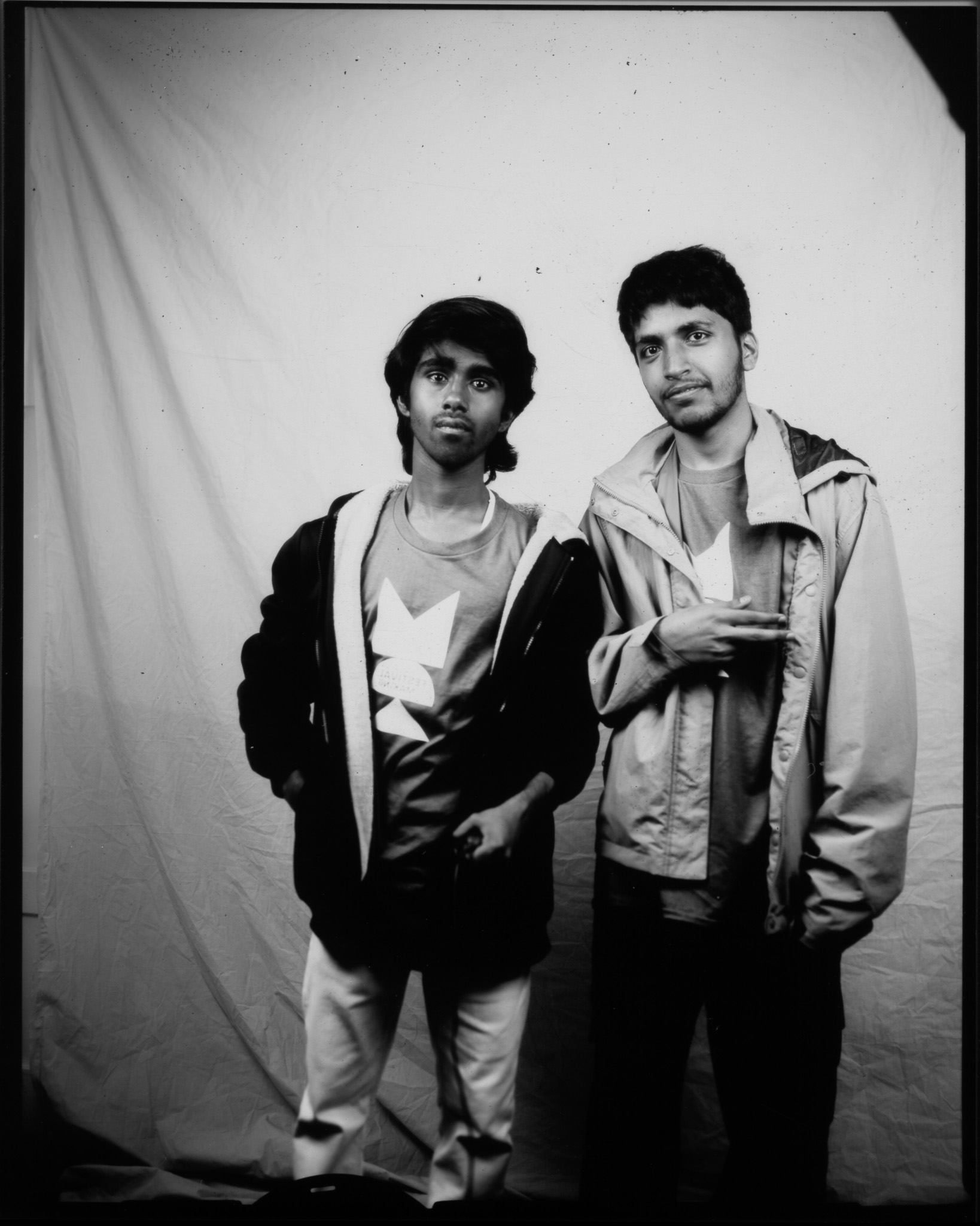 Black and white image of two adults stood smiling in front of a white sheet. One has their arm up with fingers outstretched.