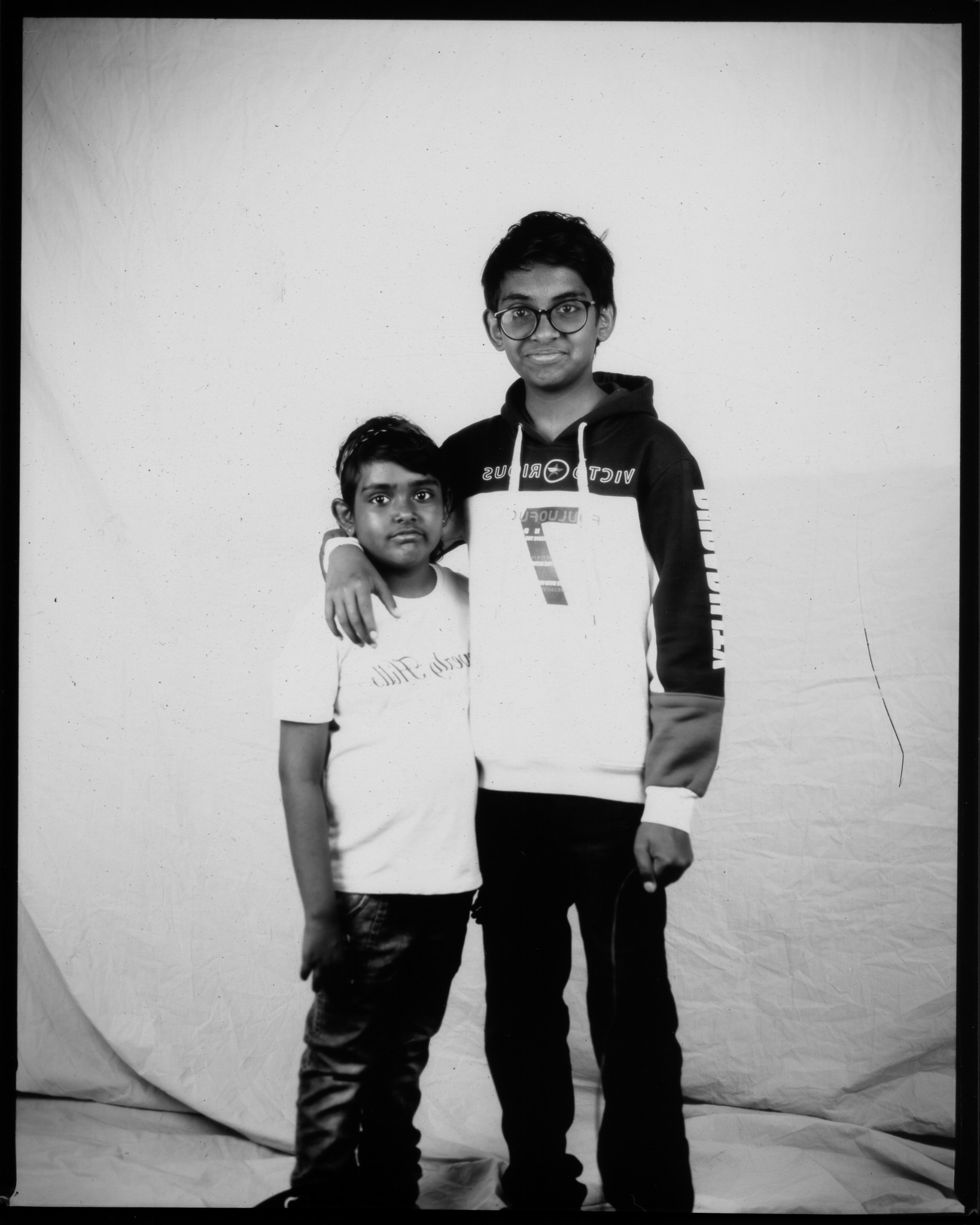 Black and white image of two smiling children, the taller child has their arm around the other's shoulder.