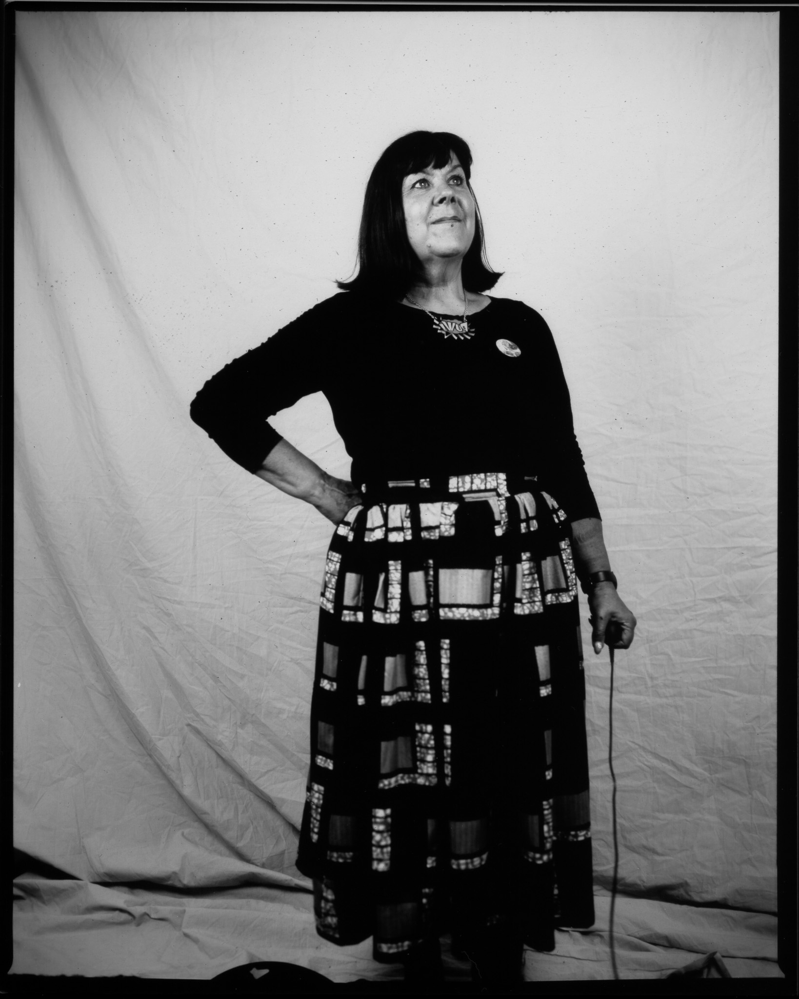 Black and white image of an adult stood looking away, a hand on their hip and the other pressing a remote with wire trailing.