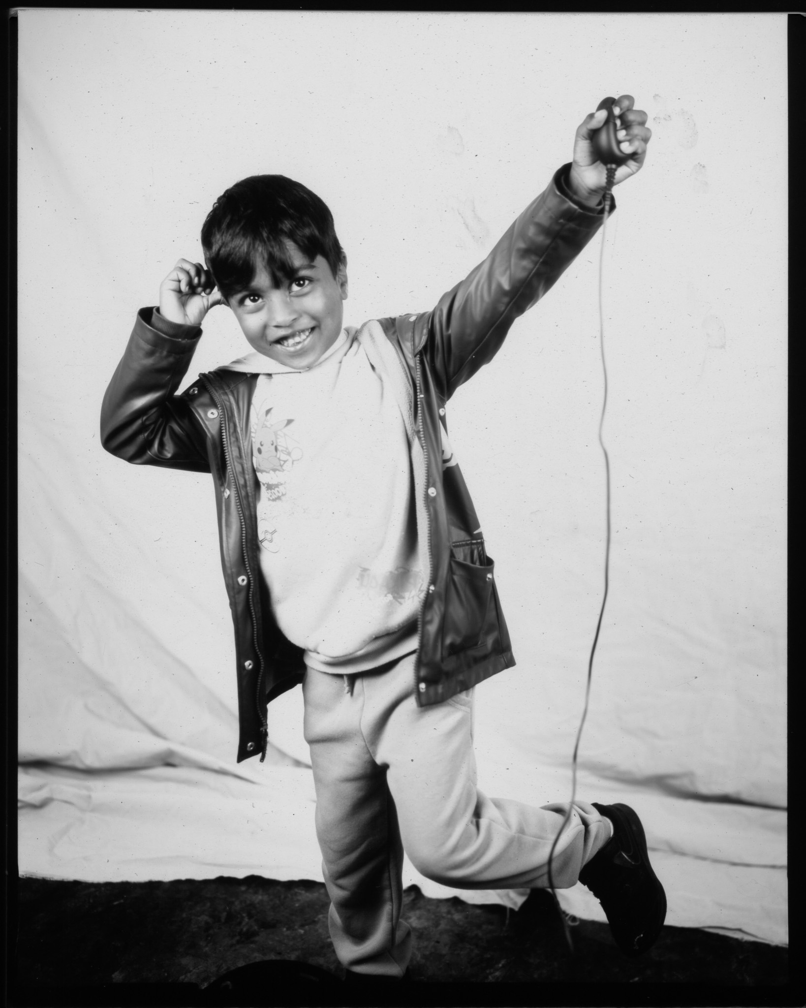 Black and white image of a smiling child with one foot lifted and arms raised. Pressing a remote with trailing wire.