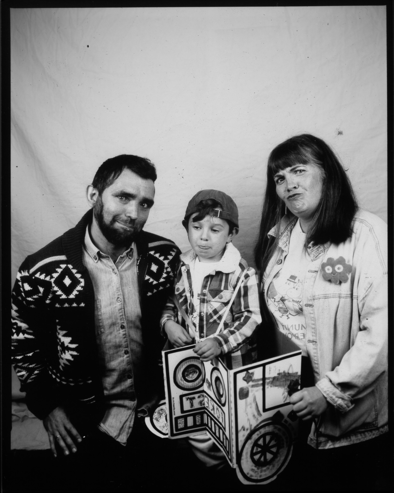 Black and white image of two adults kneeling next to a child stood in a cardboard car. Each with a different expression.