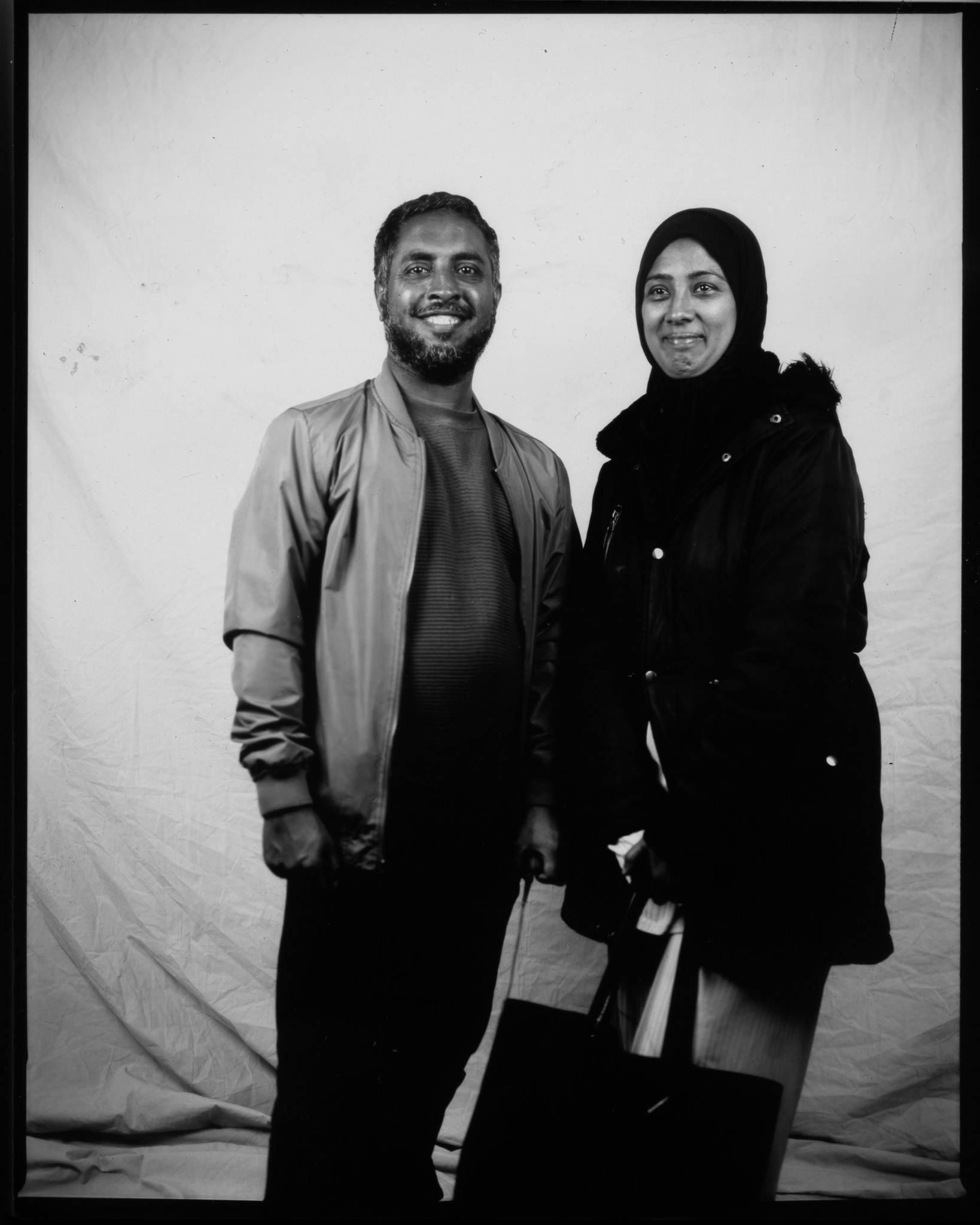 A black and white image of two adults stood smiling in front of a white sheet. One adult looks away.