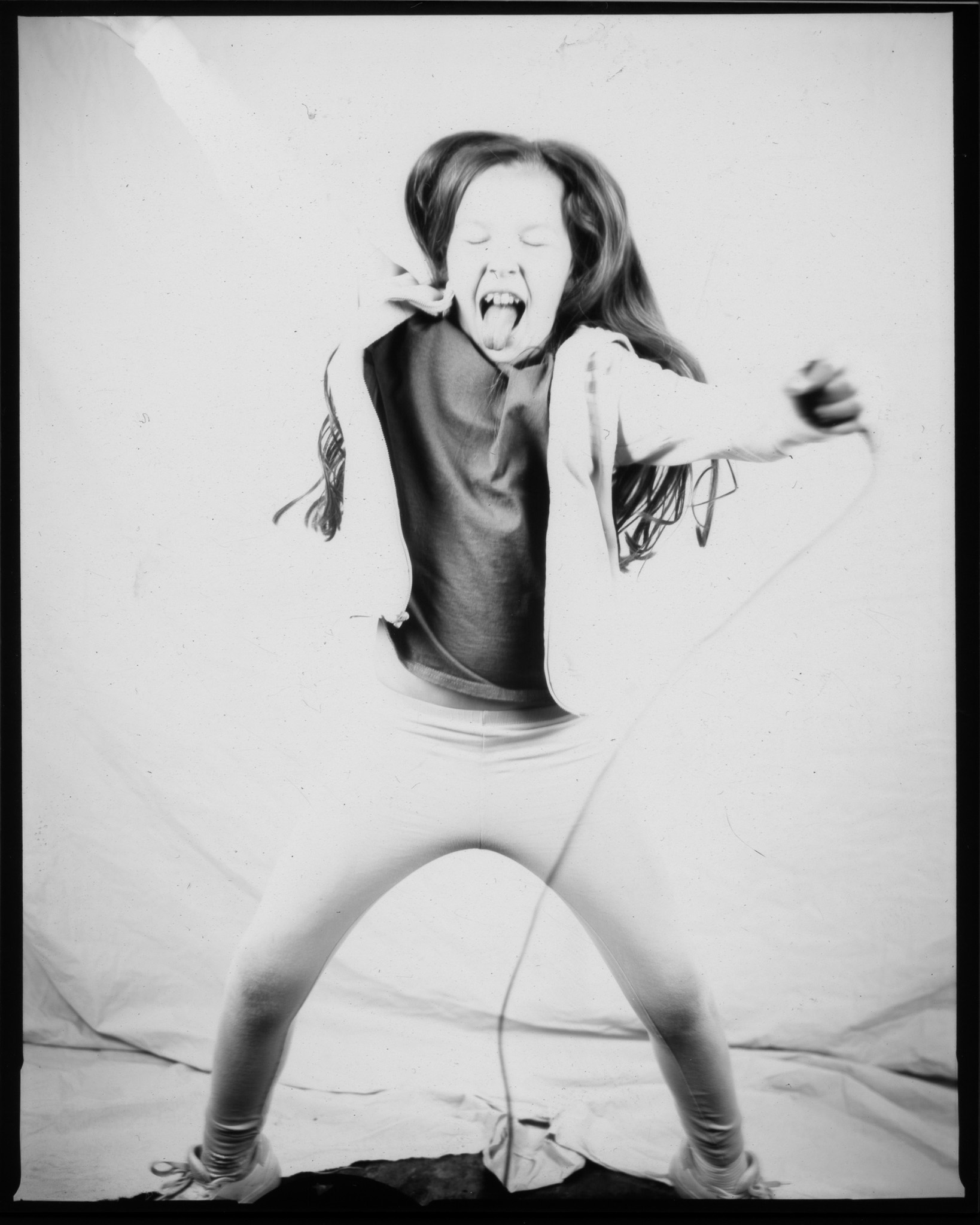 Black and white image of a child mid-star jump, tongue stuck out and pressing a remote with a wire trailing down.