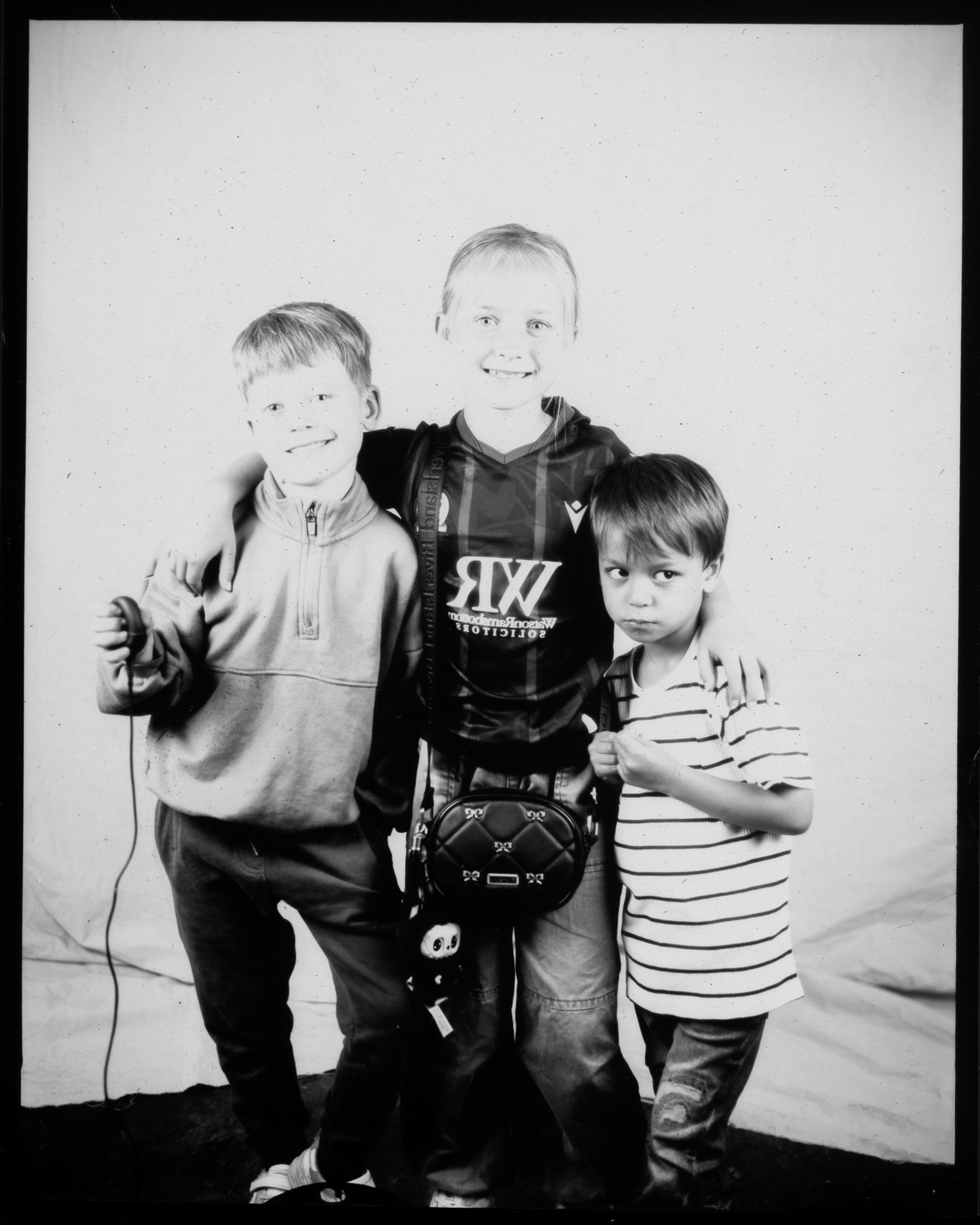 Black and white image of three children, one has their arms around the others. One child presses a remote with wire trailing.