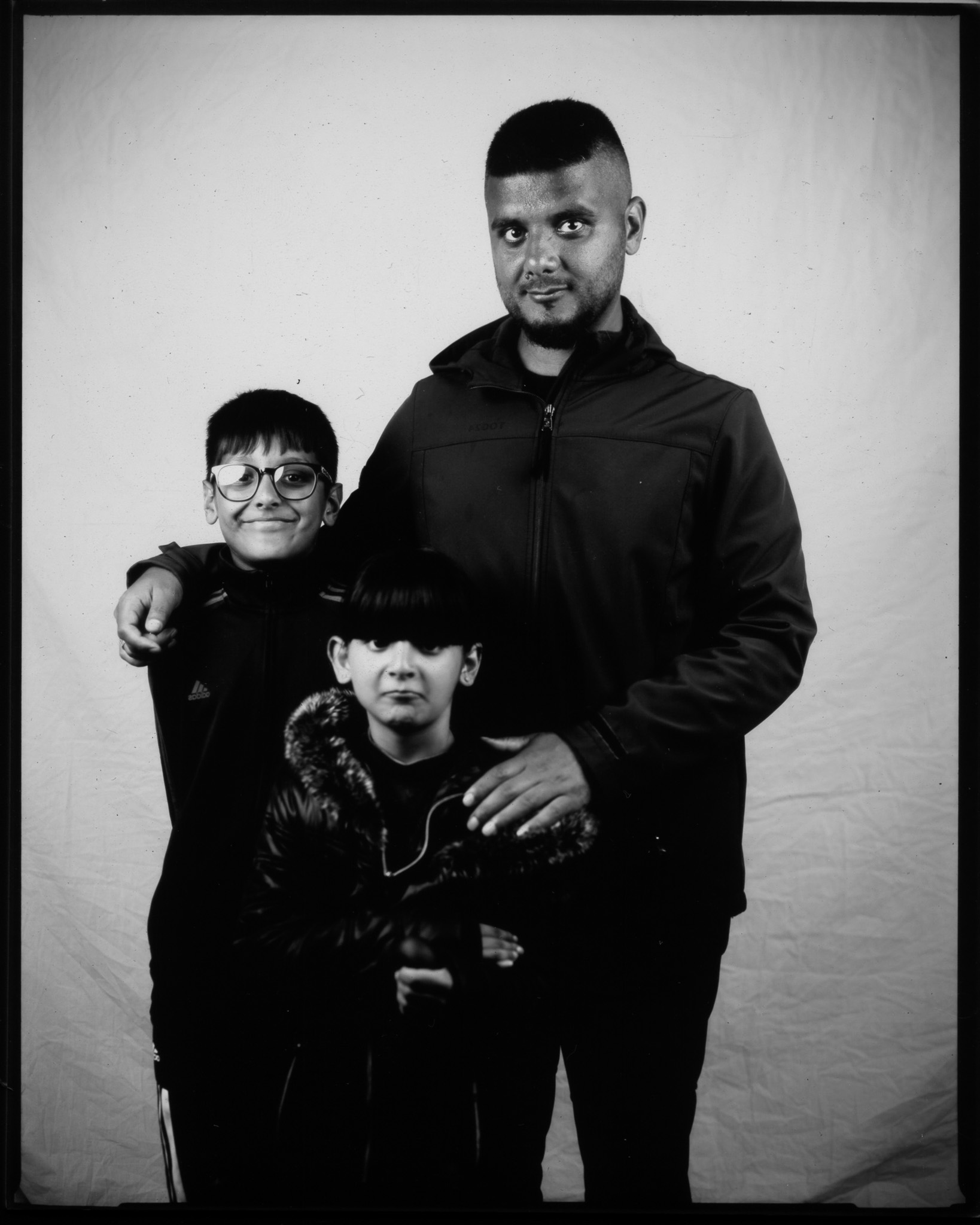 Black and white image of an adult with their arms around two children, all stood smiling in front of a white sheet.