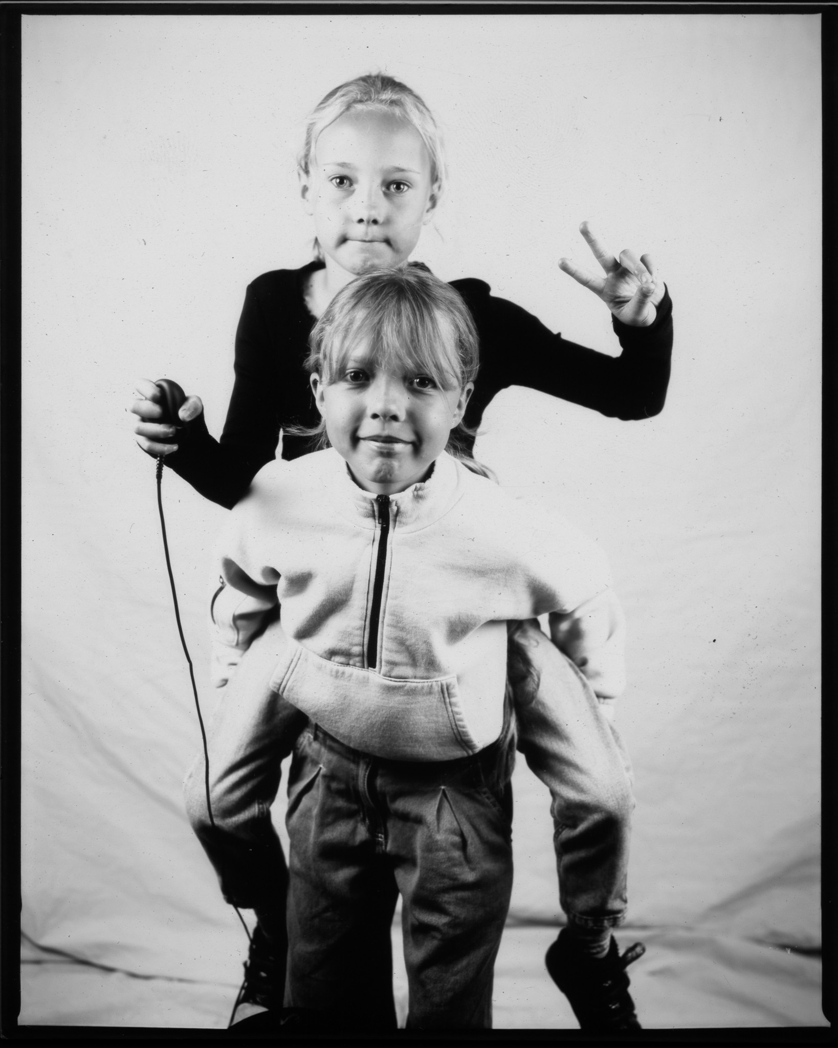 Black and white image of two children, one giving a piggy back. The child on top makes a peace sign and presses a remote.