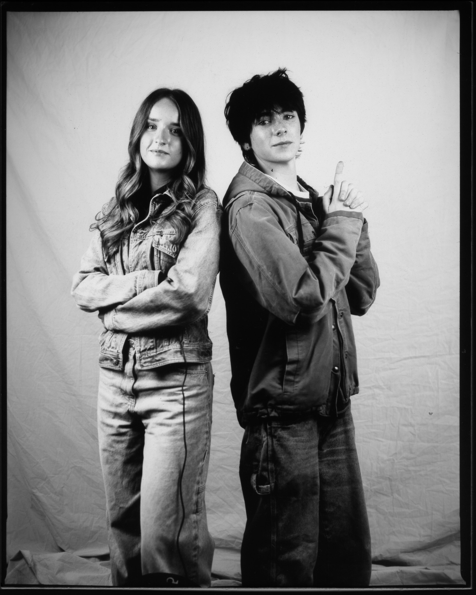 Black and white image of two teens back to back. One has arms crossed and a trailing wire, the other points up.